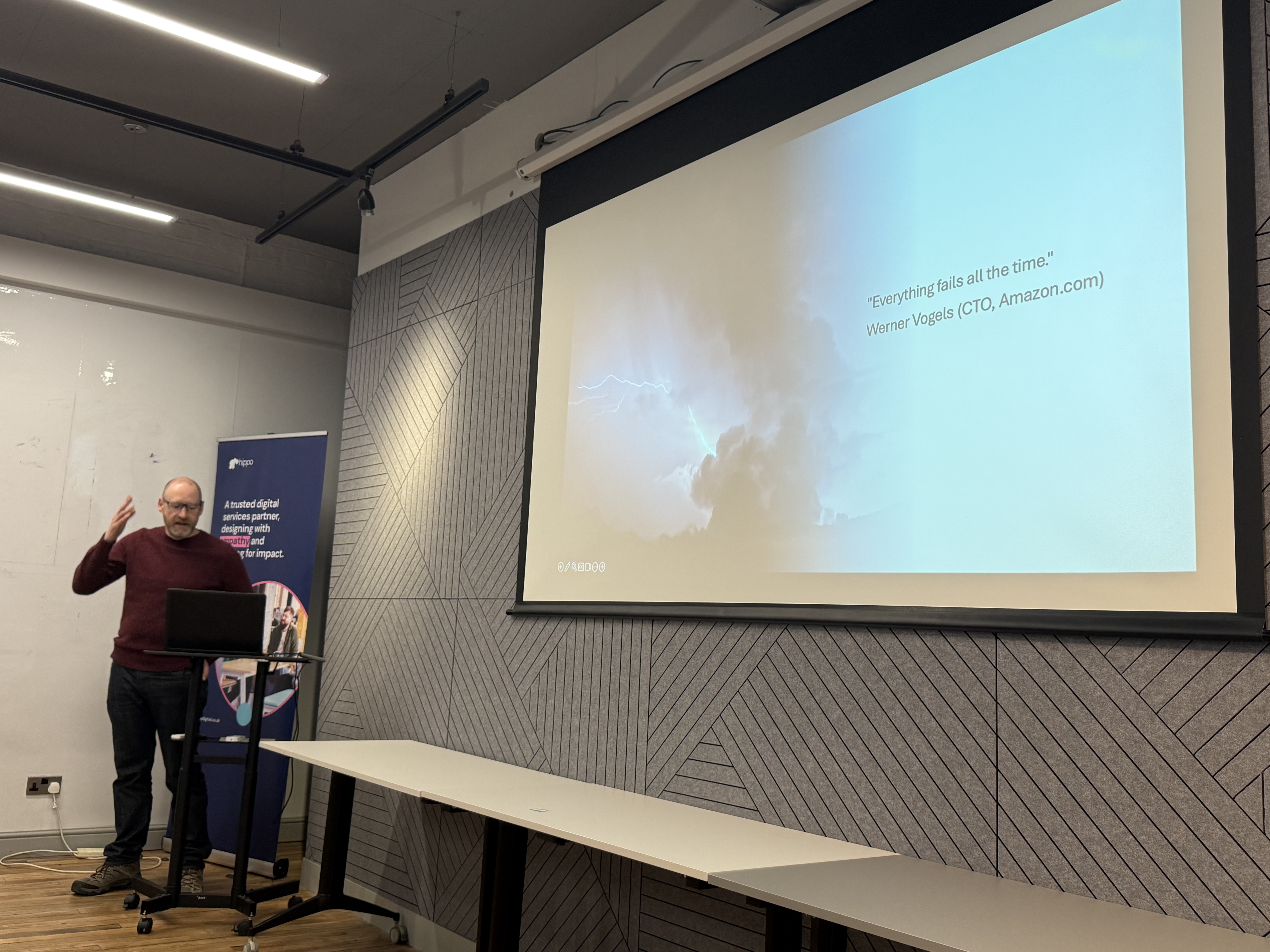 Alex, a male in black jeans and plum top who is wearing glasses and has short hair and beard. Stands at the front wit... image