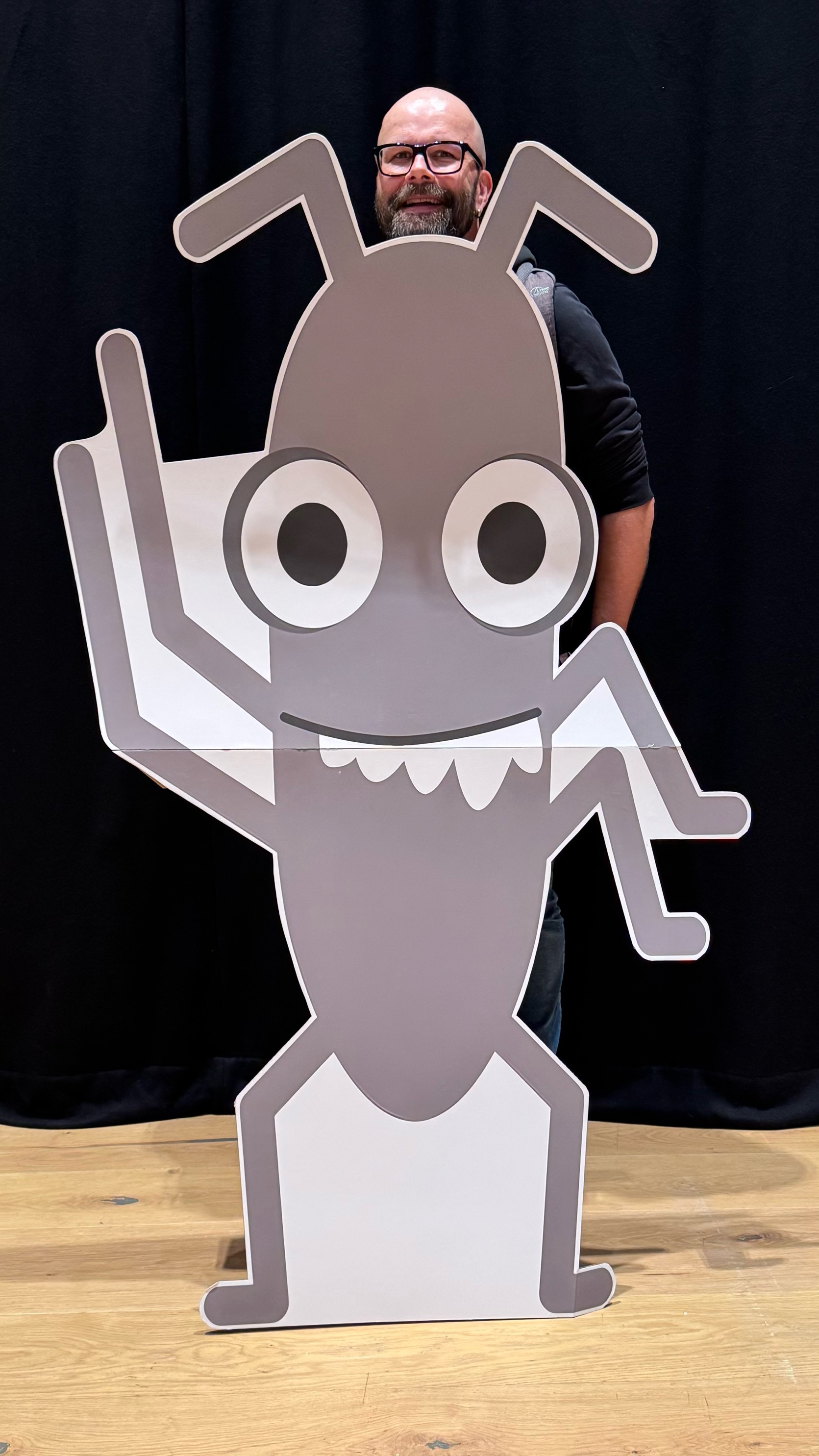 Christian peeking out from behind a cardboard cutout of the Ministry of Testing’s cheerful 'Bug' mascot, smiling with just his head visible above the character.