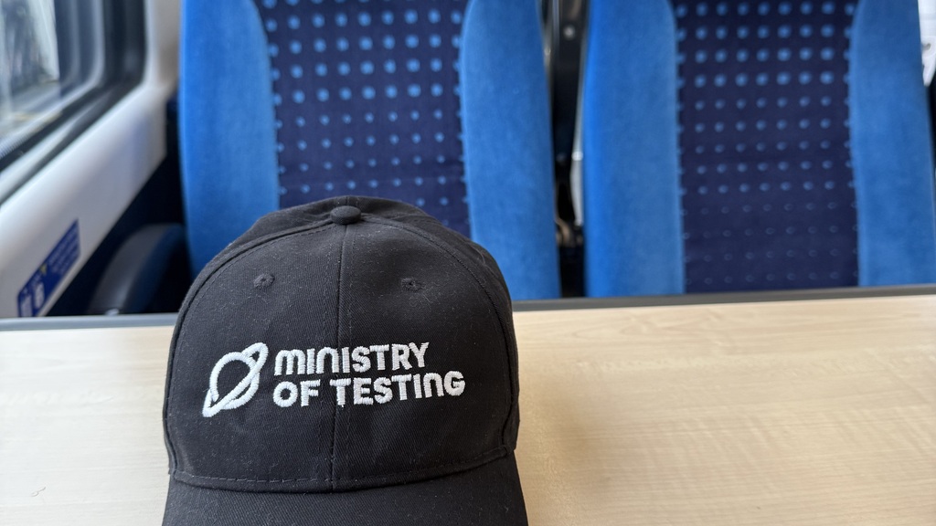 A black baseball cap with the planet logo and Ministry of Testing sits on a train table with two blue train seats in ... image