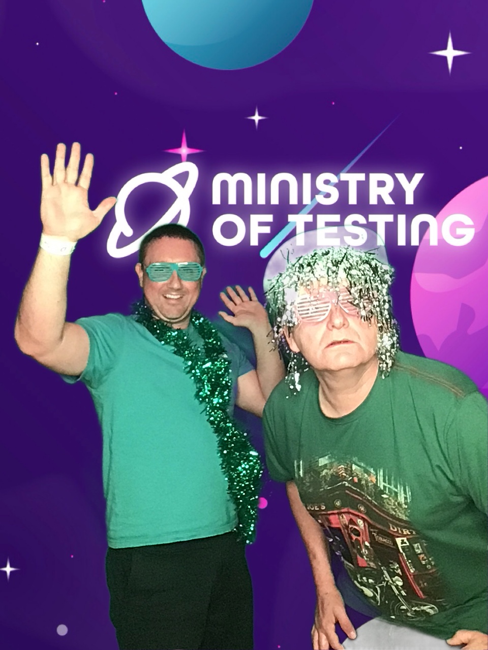 This pic shows two TestBash attendees both wearing green and decorated like a christmas trees. Attendee no1 on the left wears a green tee, green tinsel and green silly cut out glasses. They are standing to the back of the frame and have their hands up and palms facing forward with a smile on their face. Attendee no2 is also wearing a green tee but is a slightly darker green. You can just about make up the design on the front is of a classic english pub with a motorbike parked outside. Atendee no2 wears a green tinsel wig, white cut out glasses and a hat on top the wig. Attendee no2 is closer to the camera and looking straight into the camera with a curious look. This is a very fun and festive feeling photo. 

Background: The green screen backdrop depicts a simple illustrated outer space scene. The main colour is purple, with cute stars and planets in shades of pink, blue, and white. About a third of the way down the screen, a logo appears. It reads "Ministry of Testing" in bold white text with a blue shooting star behind it. Next to the text, in the same bold white style, is a line drawing of the planet Saturn.