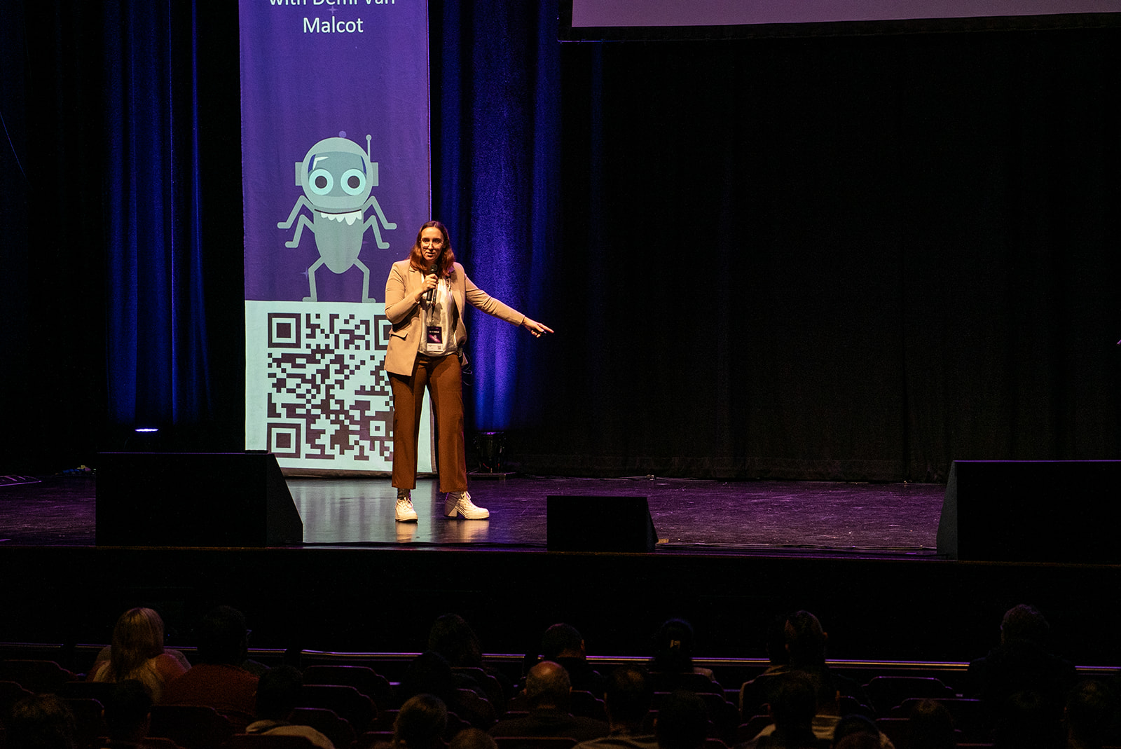 A smiling woman with light brown hair stands on a dimly lit stage in front of a large screen, speaking into a beige headset microphone. She is wearing a light-coloured blazer over a darker shirt, and rust-coloured trousers and awesome high-heel Converse trainers, pointing to her right with her left arm extended. Behind her, a purple banner on the screen features a cartoon robot icon and a large QR code below it. The stage floor is dark, and the bottom third of the image shows the tops of the heads of the audience seated in the foreground.