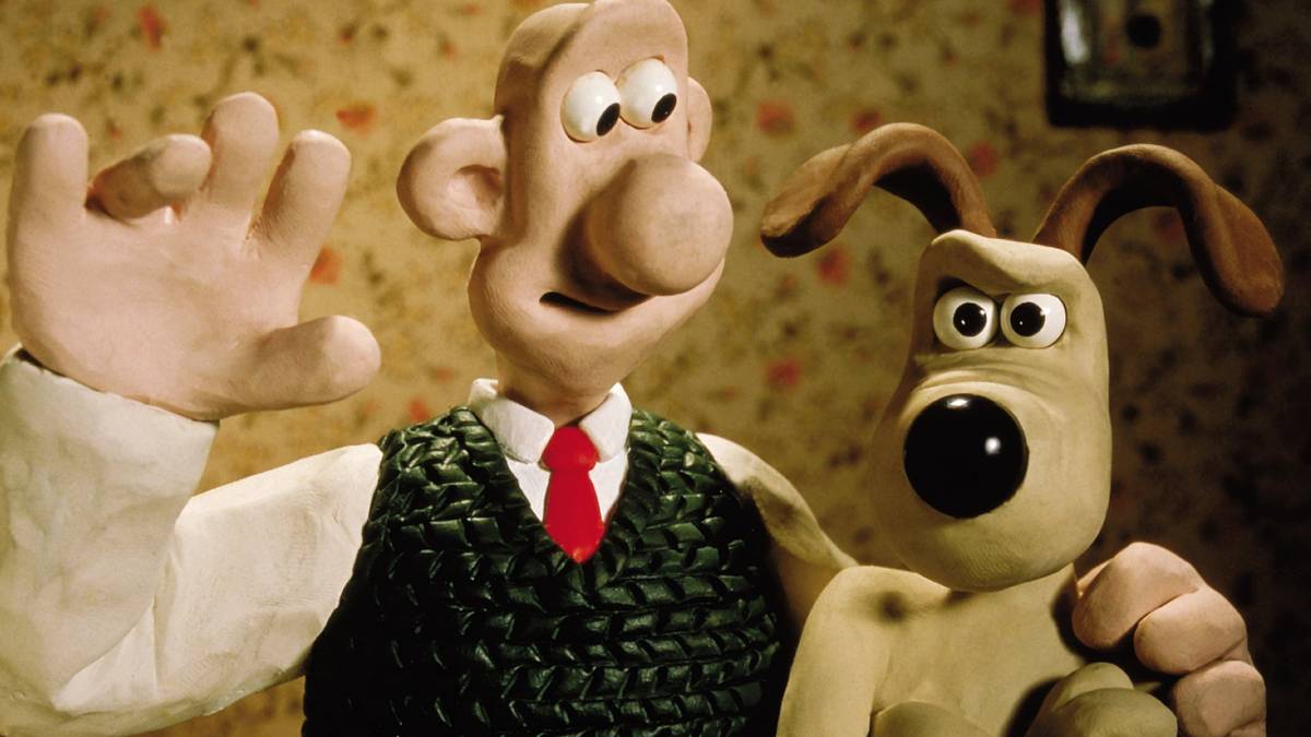 Cartoon characters Wallace and Gromit. Wallace, clad in white shirt, red tie, and green vest, is smiling and waving at the camera. He has his left arm around the shoulders of his dog Gromit, who is looking up at him with skepticism and annoyance.