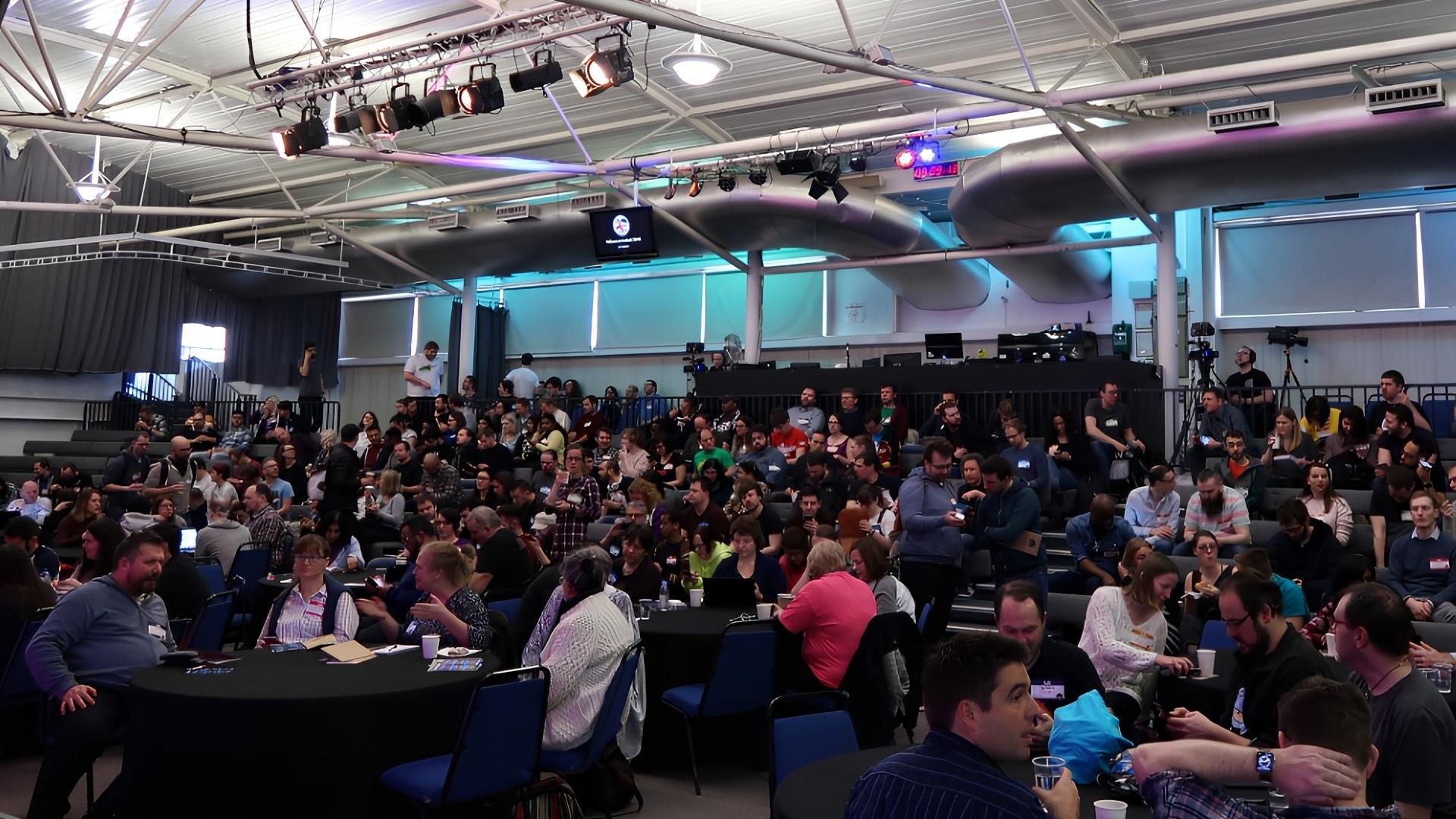 About a hundred or so people gathered in a conference space. There is an impressive lighting rig attached to the ceiling. Conversations are in full flow. There is a bug and duck character attempting to hide.