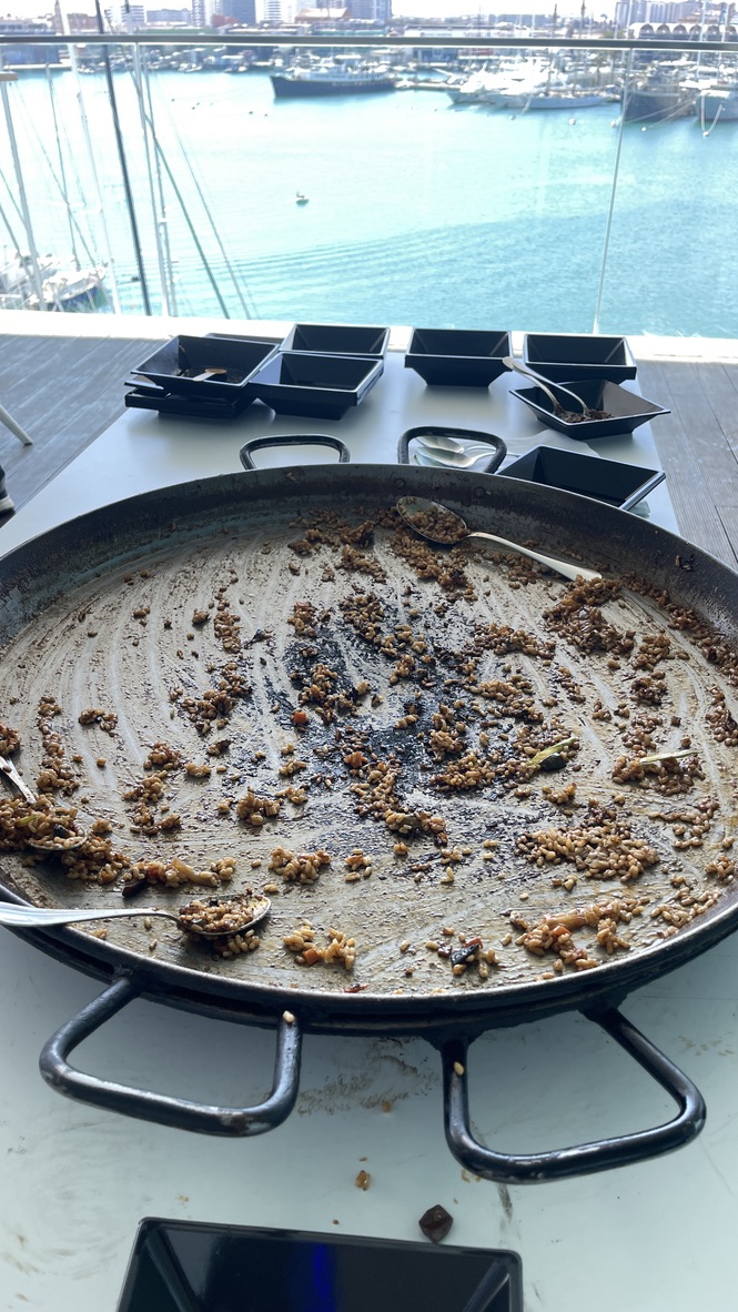 An enormous, nearly empty paella pan sits on a table overlooking the marina in Valencia, with only a few scattered grains of rice and spoons left behind — clear evidence of a well-loved lunch at Selenium Conf 2025.