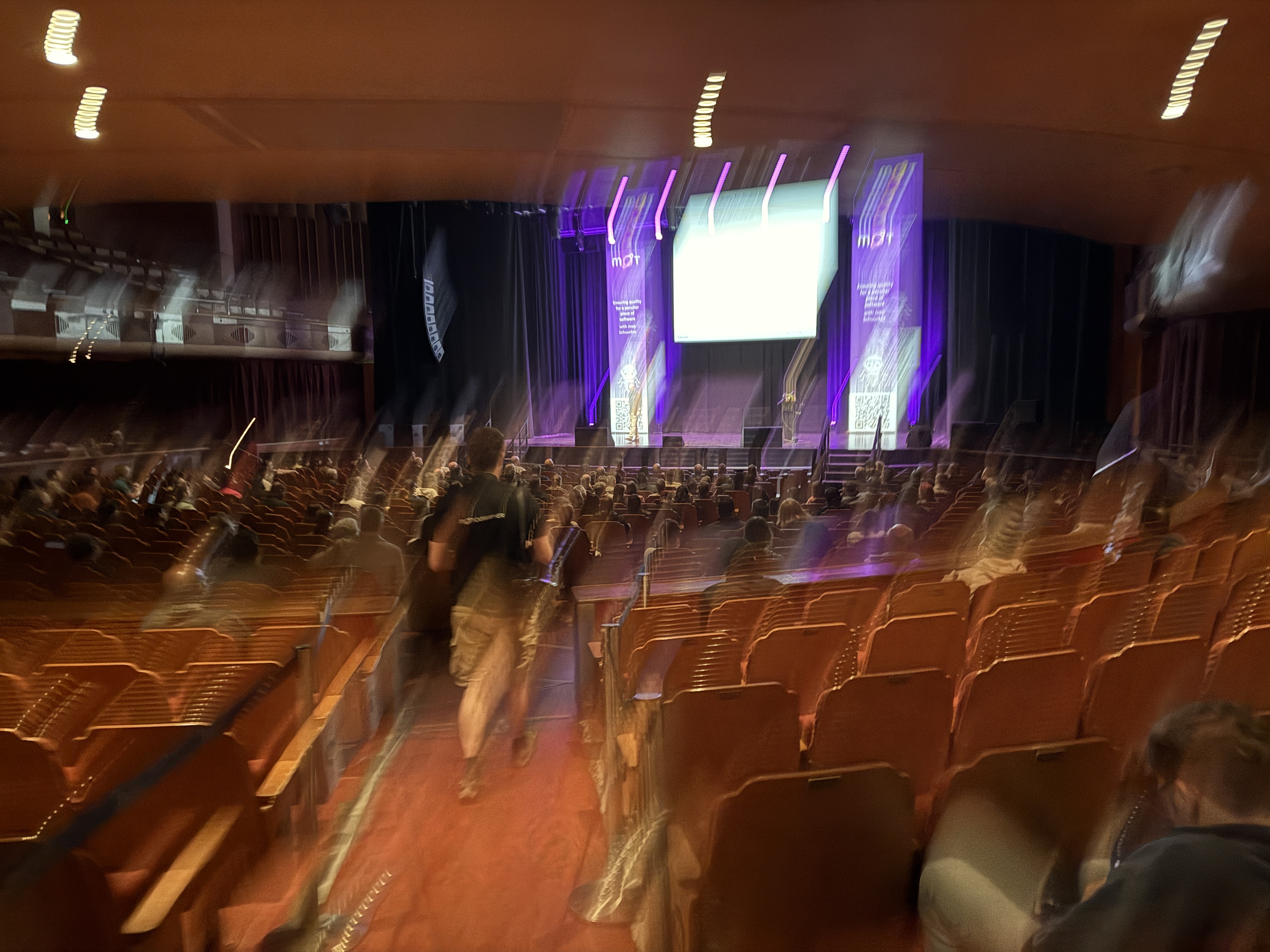 A long exposure, motion-blurred image of a large auditorium from the back. On the stage, a large bright screen is centred between two banners with a purple design and the letters 'MOT' visible. A few people are walking down the central aisle toward the stage, and the motion blur emphasises their movement.
