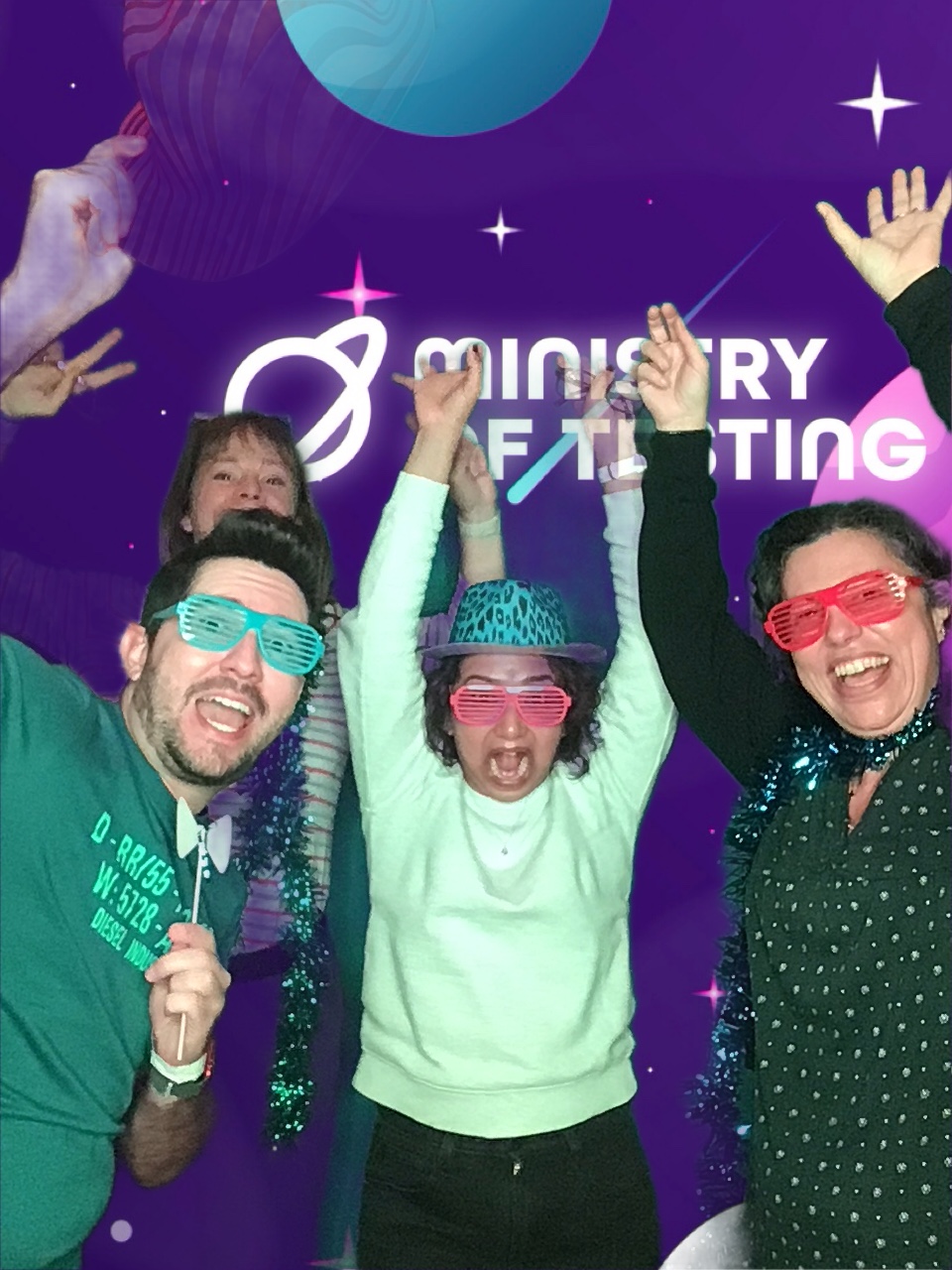 Here we have the rollercoaster pose as a group of 4 attendees at TestBash 2024. all attendees have both arms in the air and are expression glee, excitement and feigned terror for their imaginary rollercoaster. Attendee 1 on the left is wearing a green tee with some writing on it, they also wear green cut out glasses and hold a bow tie on a stick. Attendee no2 is a little more at the back and we can jst about make out that they are wearing green tinsel around their neck along with a stripey long sleeved top. Attendee no3 is smack bam in the middle and wearing a lime green sweater matched perfectly with a green leopard print hat and orange cut out glasses. Attendee no3 has a large expression of the rollercoaster pose, with both their arms stretched out as high as they can go and their feign facial expression of terror is maxed out too. Attendee no4 has both hands up in the air and look like they are really enjoying themselves. They have tinsel around their neck and orange cut out glasses on their face. 


Background: The green screen backdrop depicts a simple illustrated outer space scene. The main colour is purple, with cute stars and planets in shades of pink, blue, and white. About a third of the way down the screen, a logo appears. It reads "Ministry of Testing" in bold white text with a blue shooting star behind it. Next to the text, in the same bold white style, is a line drawing of the planet Saturn.