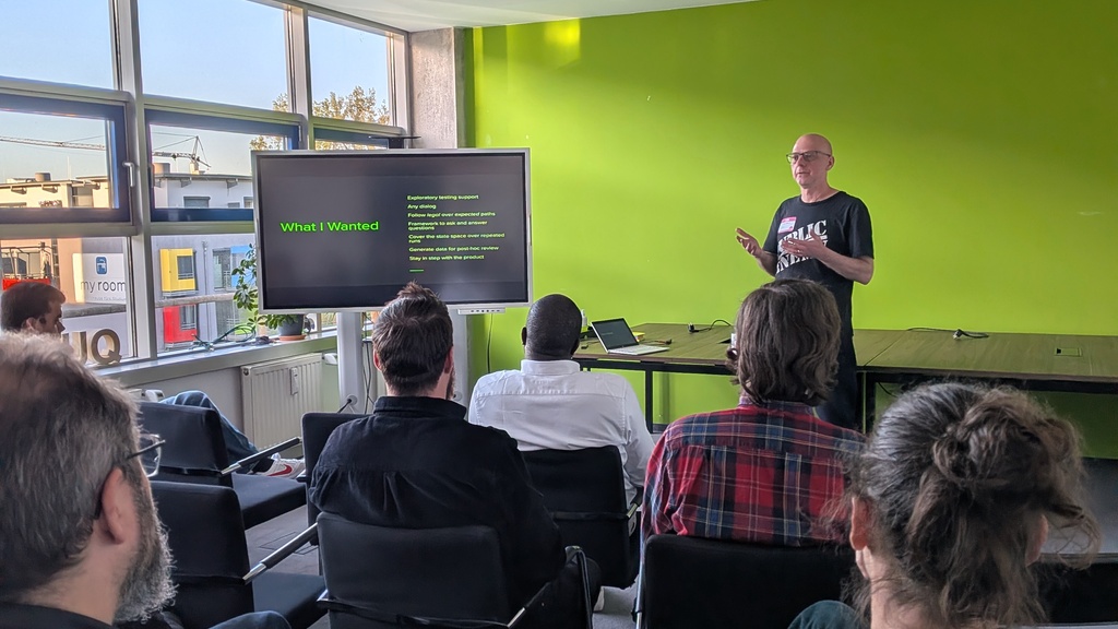 A person gives a presentation to a seated audience in a room with a green wall and large windows, displaying slides o... image