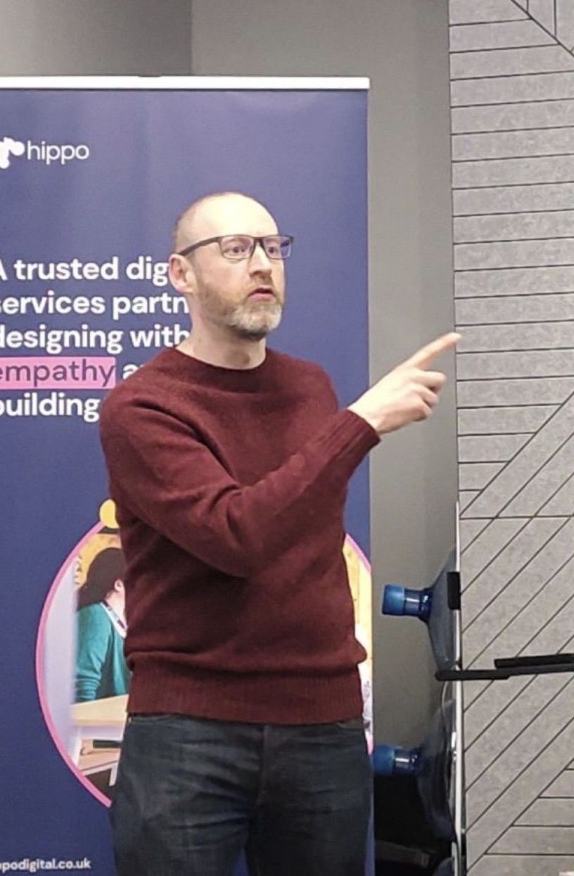 Alex is a white male in black jeans and a plum top, wearing glasses. He is stood speaking and pointing in front of a ... image