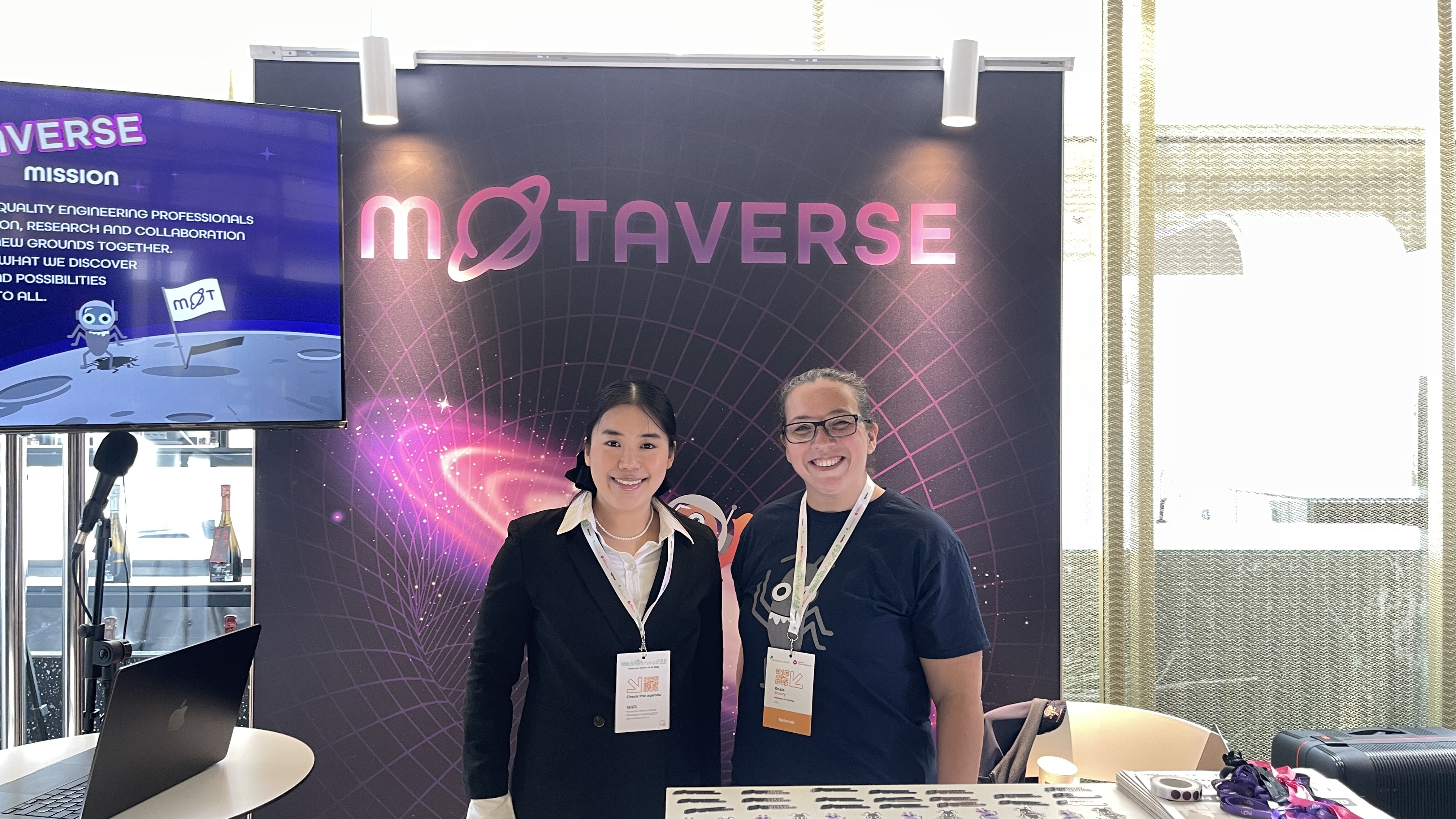Amalie Shi stands beside Rosie Sherry at the Ministry of Testing stand after her talk at Selenium Conf 2025. They’re smiling in front of the MoTaverse backdrop, with stickers and materials on the desk in front of them.