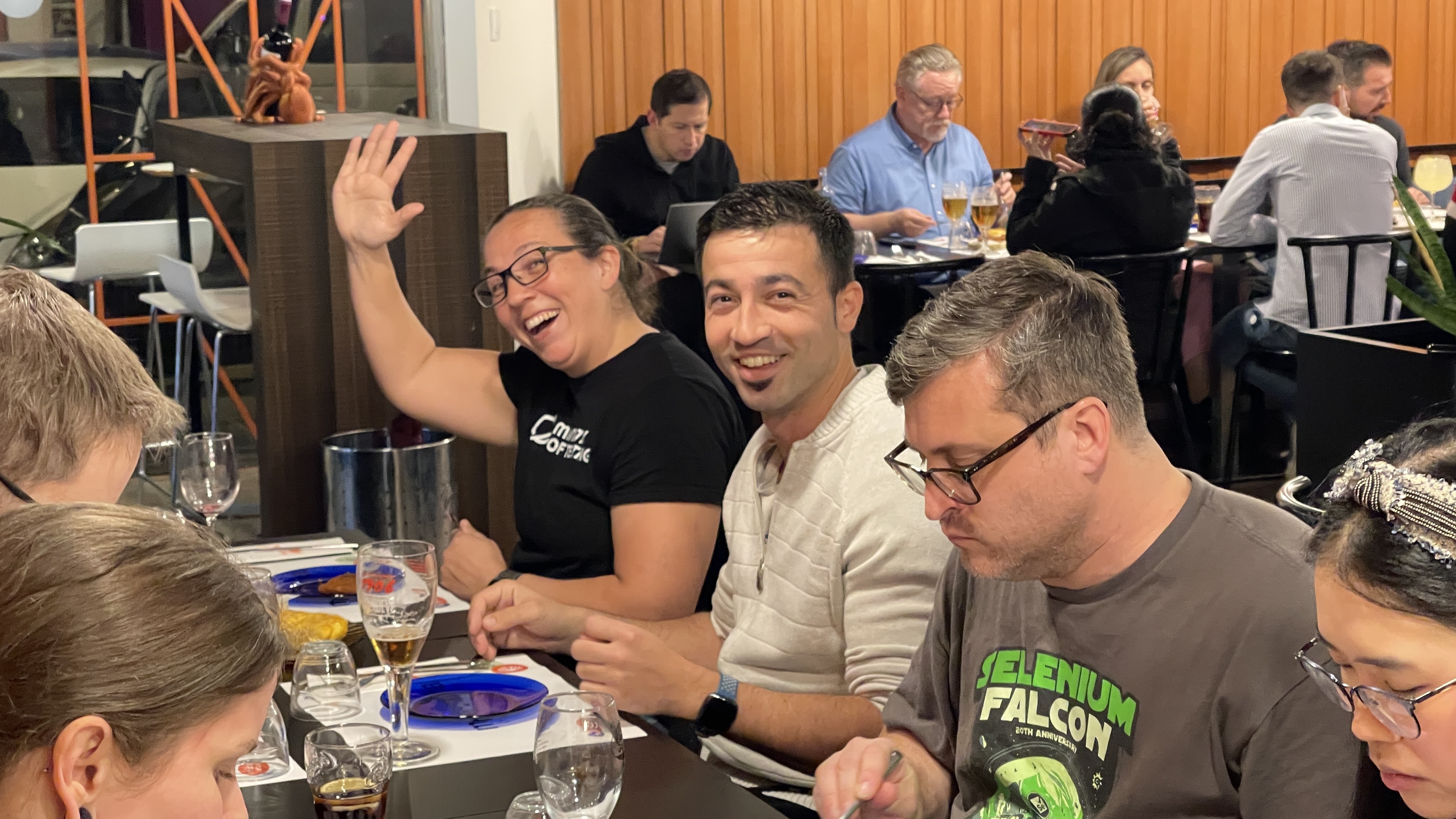 Rosie Sherry waves with a big smile beside Sergio Olmos, Titus Fortner and Amalie Shi at the Selenium Conf 2025 speakers' dinner at Maruxiña in Valencia. In the background, Diego Molina is working on a laptop, and Merrill is enjoying food at another table. Glasses and plates of Galician cuisine are spread across the table.