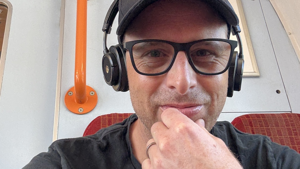 A selfie of a middle aged man with a black t-shirt and cap sat on a train. 