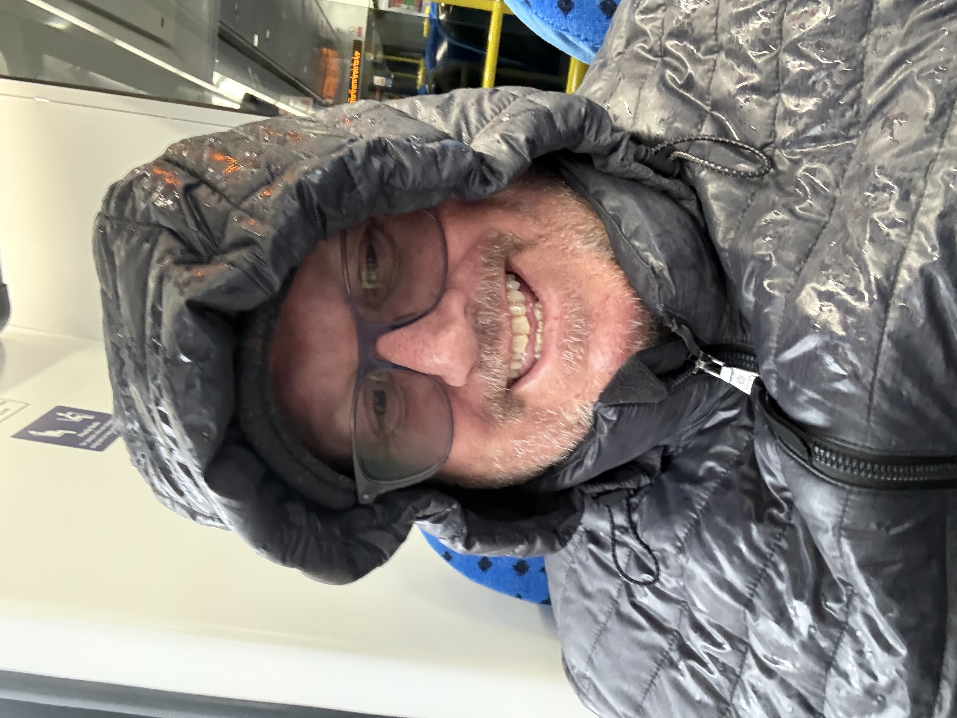 Selfie of Ady cap on, hood up, and looking very wet sat on the train. 