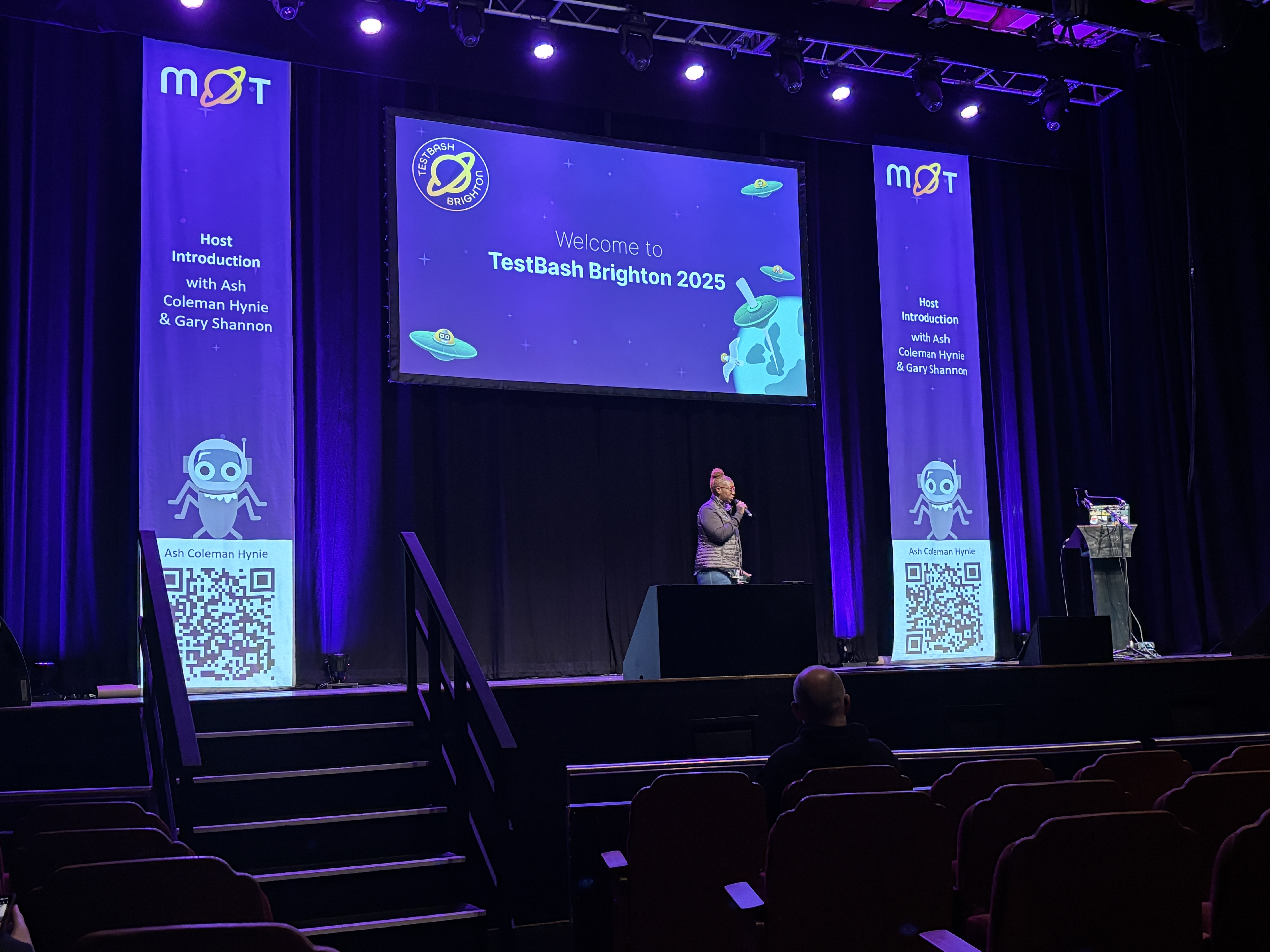 Here's the alt text for the photo:
A person standing on a stage at a conference, speaking into a microphone. The large screen behind them displays the text, "Welcome to TestBash Brighton 2025," with a space-themed graphic featuring a robot and flying saucers. Two tall vertical banners flank the screen, each reading "Host Introduction with Ash Coleman Hynie &amp; Gary Shannon" and featuring a QR code below an illustration of a robot. The stage is lit with purple and white lights. In the foreground, the seating area is visible, with dark red chairs and one person seated facing the stage.
