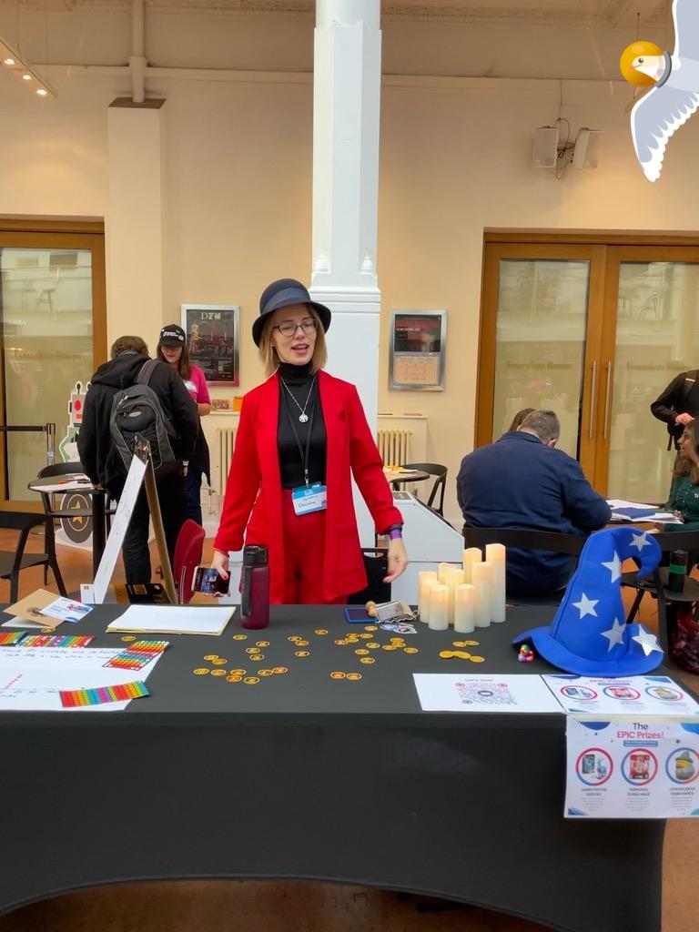 Christine Pinto is wearing her red uniform, standing behind a table in an event space. A bug, a duck and a seagull character have been added to the photo. 