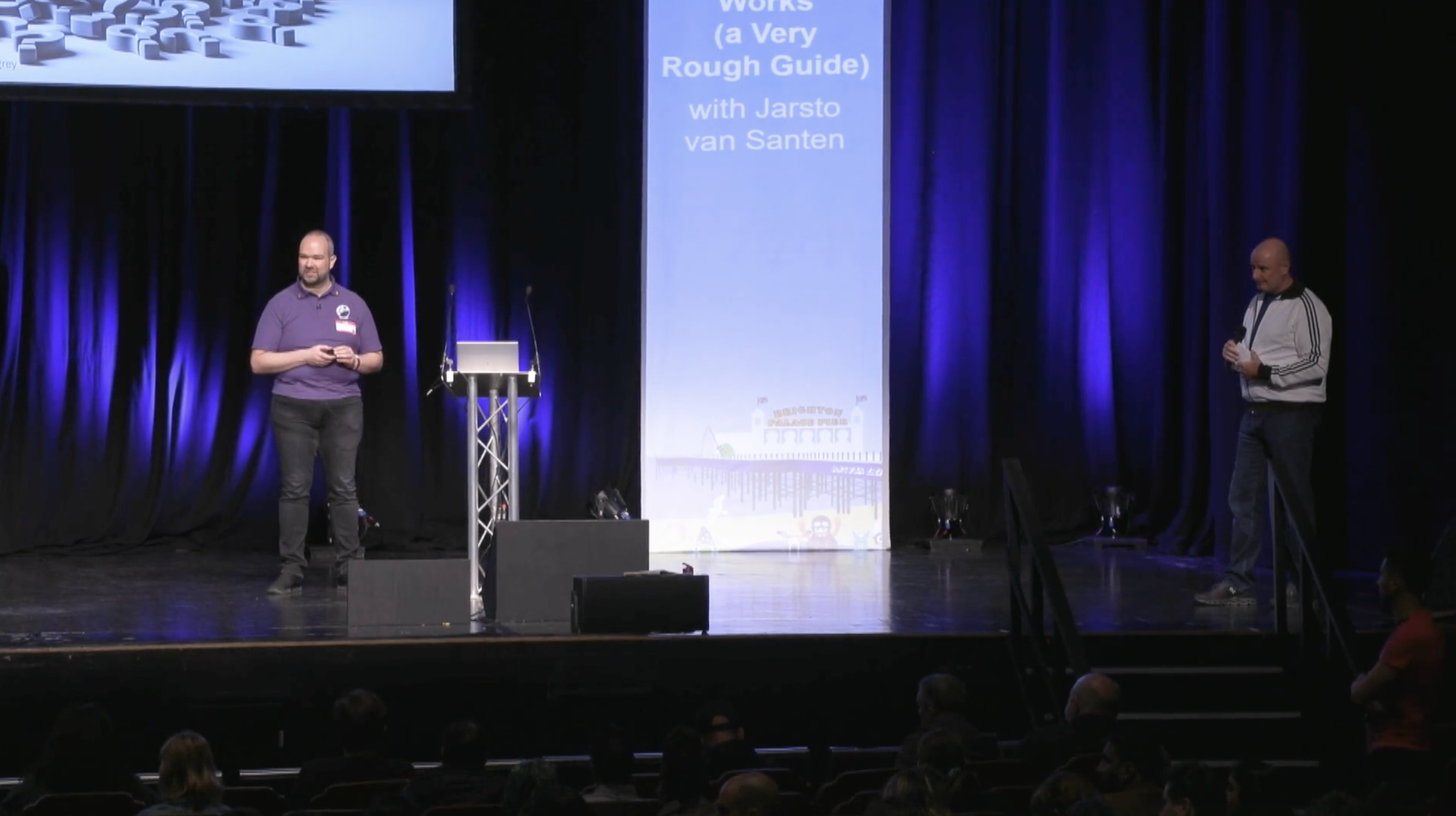 One man is presenting on a large stage. Behind him, there is a huge screen. A host is standing to the right of the stage.