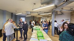 A large group of people conversing, eating and drinking in a room.

There is a long table with green pizza boxes and paper plates  image
