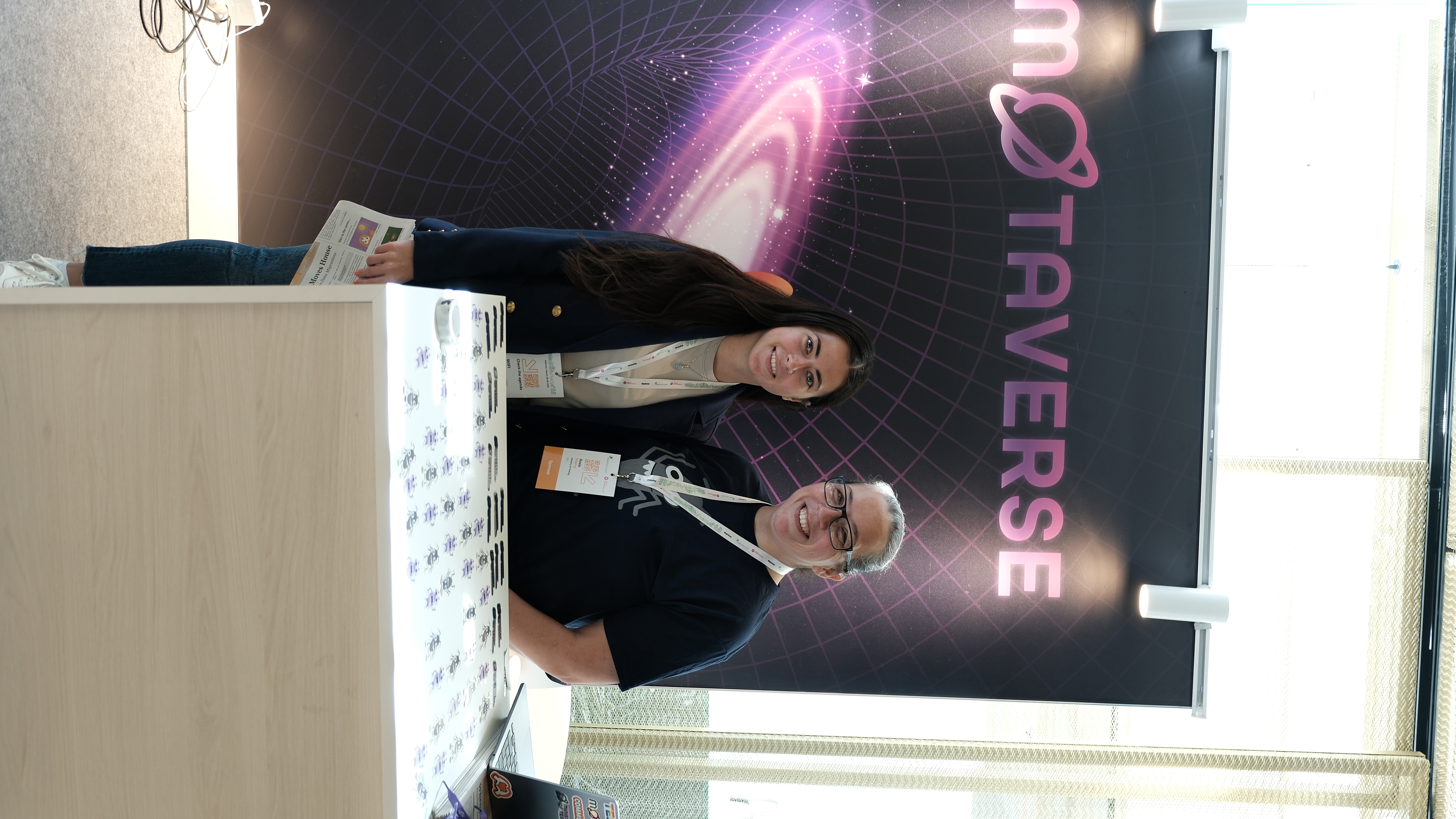 Rosie Sherry stands with an attendee at the Ministry of Testing stand at Selenium Conf 2025, both smiling in front of the vibrant MoTaverse backdrop covered in space-themed design.

