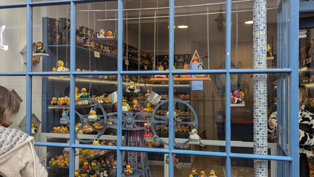 
A full exterior shot of "The Duck House" storefront, showcasing its large display window filled with hundreds of novelty rubber ducks. A person with brown hair and a cream-colored sweater is visible from the back on the left, looking into the window. The ducks are arranged on multiple shelves and platforms, varying in size, color, and theme, including ducks dressed as different characters or with various accessories. The building's facade is painted a muted blue, with white trim around the windowpanes. A narrow, vertically tiled column with a mosaic pattern is visible on the right side of the window. Part of the shop's name, "THE DUCK HOUSE," is visible at the top, along with the web address "duckhousebrighton.online." A bug, duck and seagull character have been added to the image. 