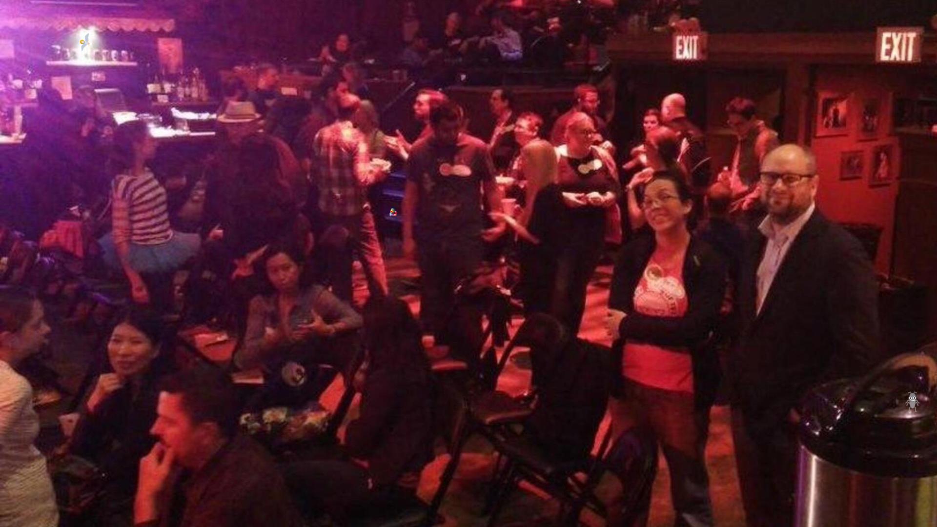 A diverse group of adults socializes in a dimly lit bar or club, which has an "EXIT" sign visible. Some people are seated at tables and chairs, while others stand and mingle. In the foreground, a woman with dark hair and glasses smiles at the camera, standing next to a man with a beard and glasses who is also looking at the camera. A bug, seagull and duck character have been added to the image.