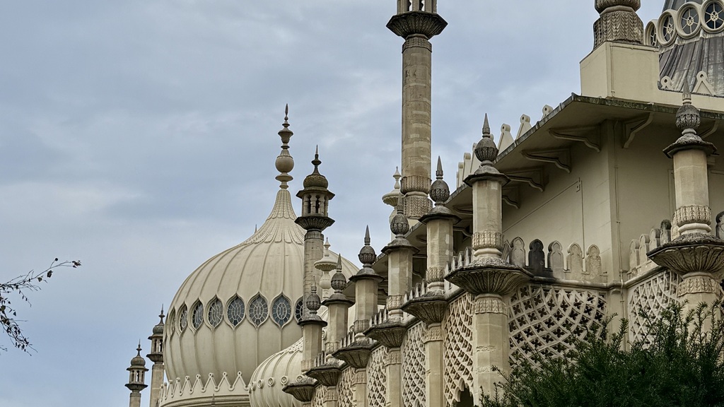 Low-angle shot of the Brighton Pavilion's elaborate Indo-Saracenic architecture, featuring minaret-like towers, a large onion dome with arched windows, intricate stone tracery, and dark green foliage in the foreground, all under a cloudy sky.