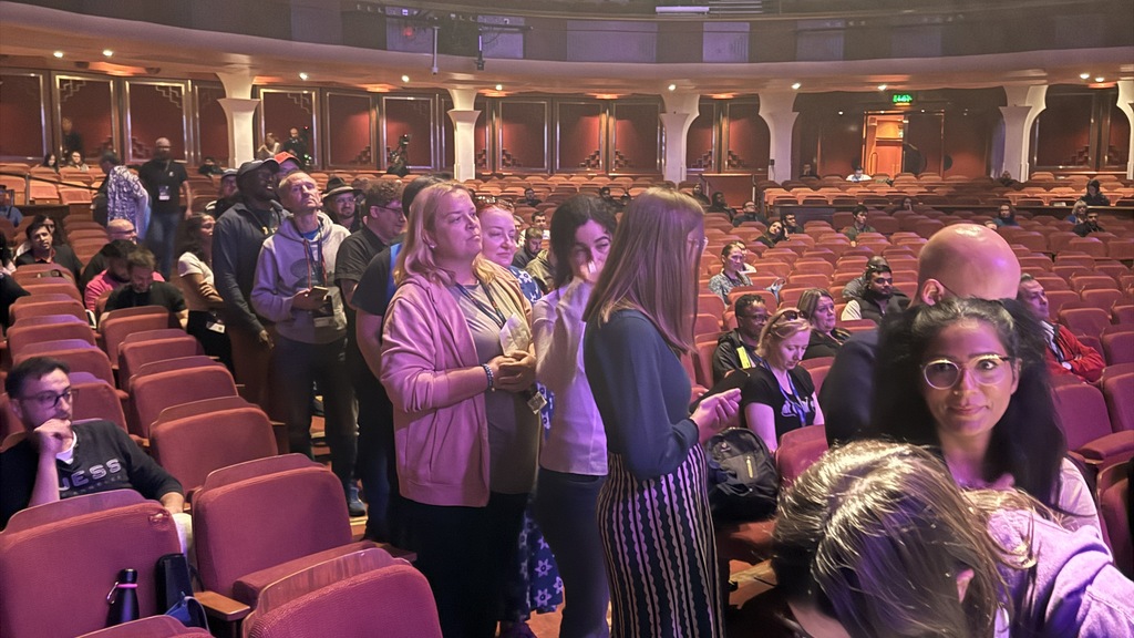 Audience members stand and mill about in the aisles of a large, ornate theatre with red velvet seats before joining the stage (out of shot). 
