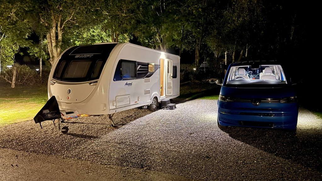 A white Swift Accord caravan and a blue VW Multivan parked side by side on a gravel pitch at night, lit by floodlight... image