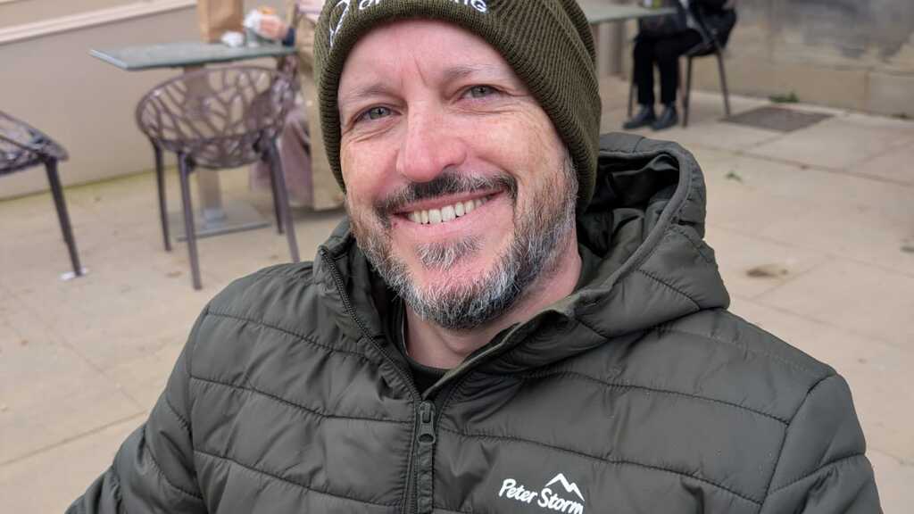 White bearded male sitting down smiling wearing a green ministry of testing beanie and a green coat image