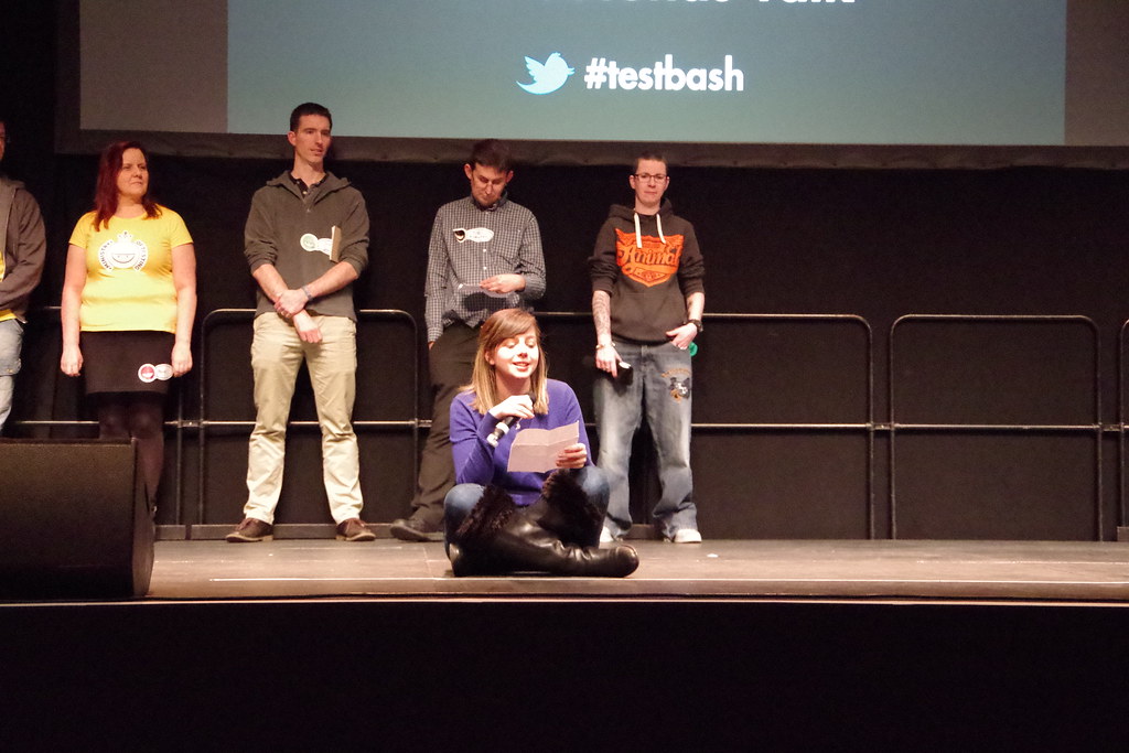 Deborah Reid on stage, sitting down, reading from a piece of paper for a 99 second talk. Behind her are people waitin... image
