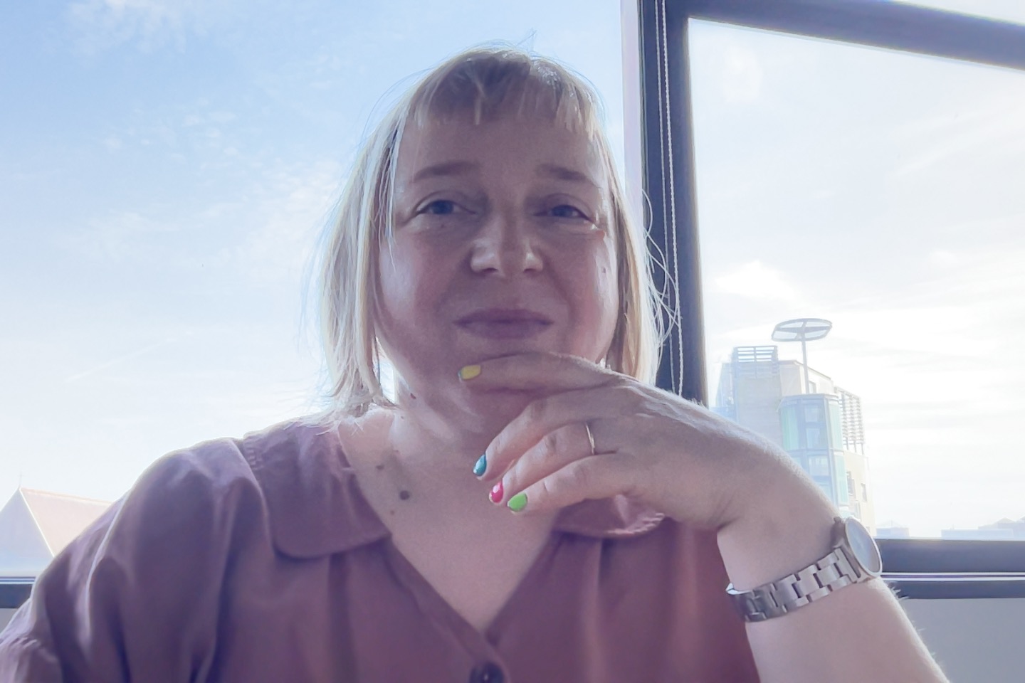 selfie of blond woman with colourful nails in front of a window with a view of sky and city