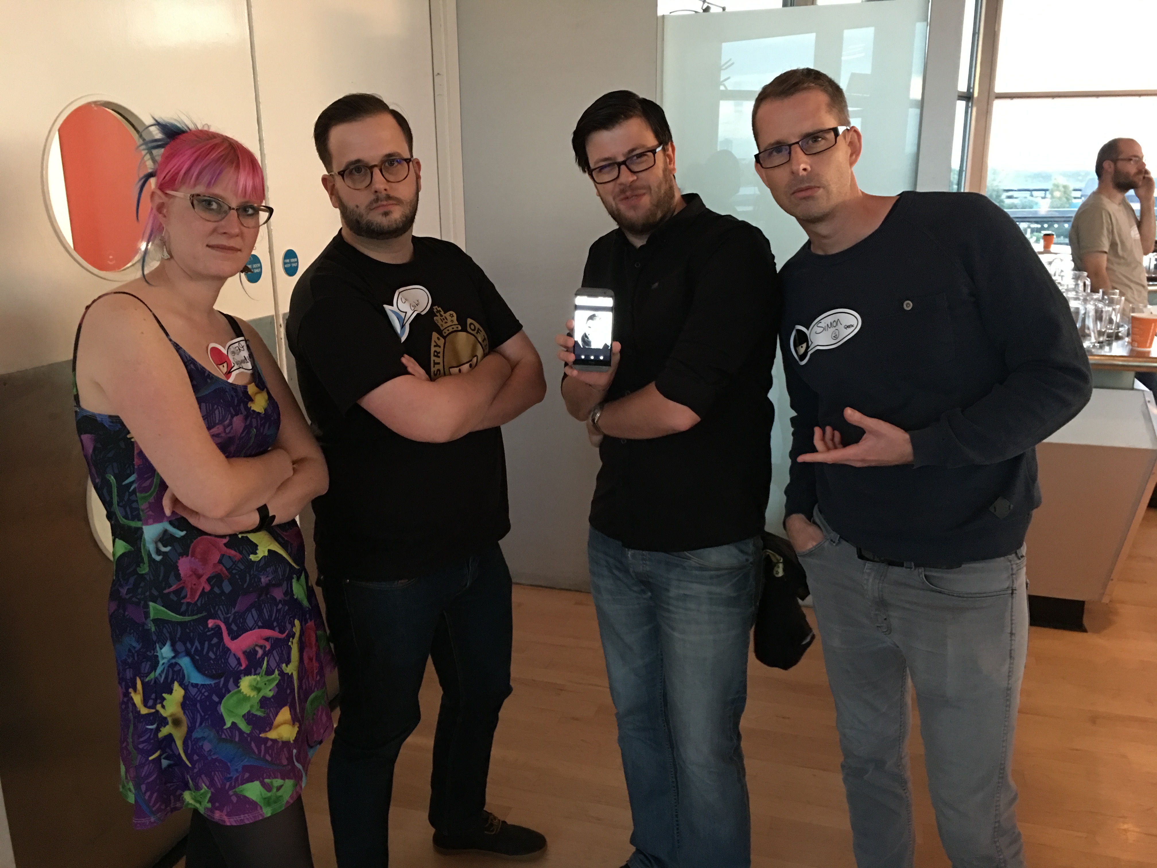 Four people. A woman and three men standing next to each other looking cool at the camera – they are attempting to look like they are in a band. One of them is holding a phone with a picture of another man.