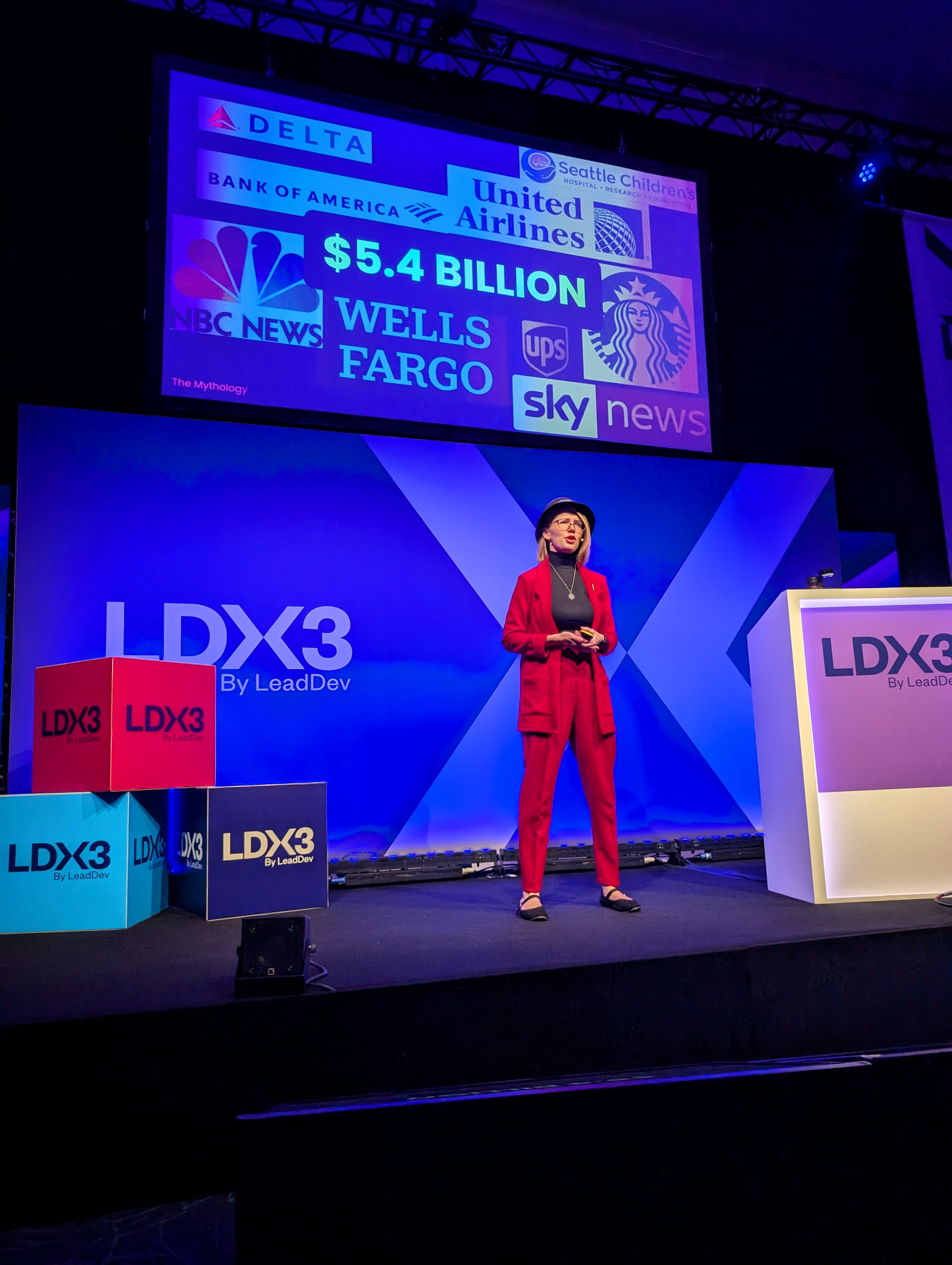 Christine standing on the LDX3 Stage. She is wearing a red suit and blue hat. Behind her is her slide showing the number '5.6 billion' and showing a bunch of logos of fortune 500 companies which lost in total 5.6 billion dollars through the global outage caused by Crowdstrike