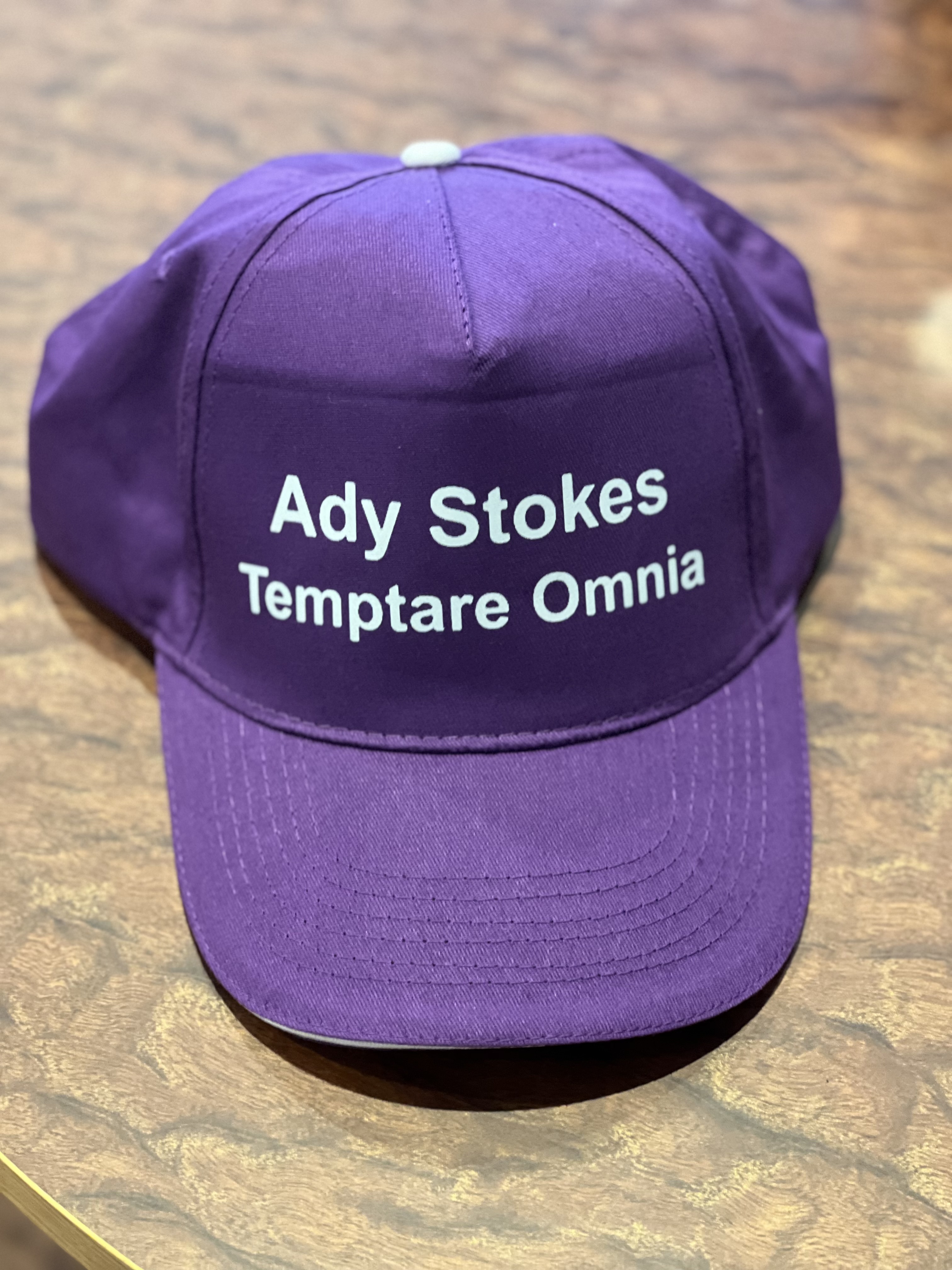A purple baseball cap on a table top. It says Ady Stokes and Temptare Omnia in white text. 