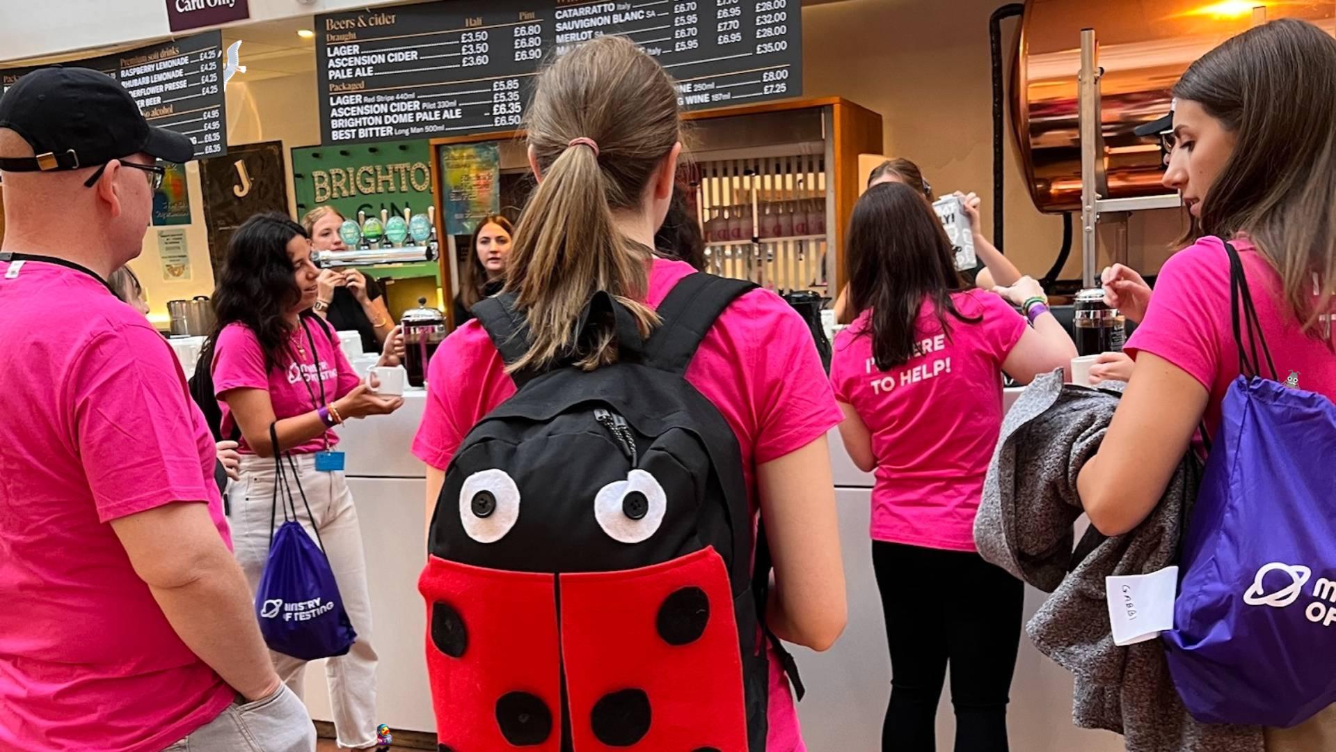 A small group of people queue up for a coffees and teas at a bar area in a conference space. They are wearing matching pink Ministry of Testing I'm Here to Help T-shirts. Some have purple Ministry of Testing drawstring bags. The woman in the middle is facing away from the camera and has a large ladybug bag on her back. A duck, seagull and bug character have been added to the photo. 