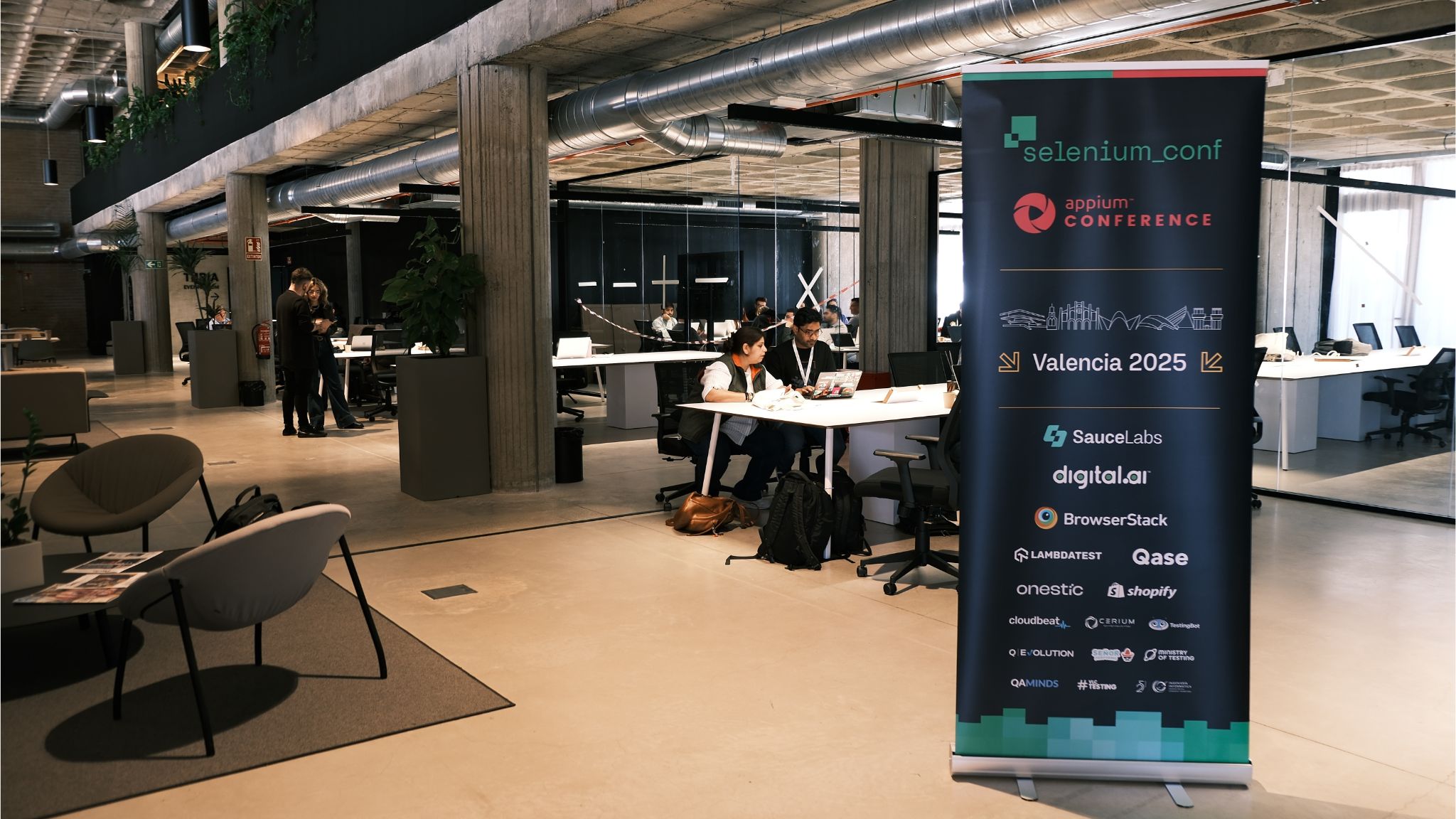 A modern, open coworking-style workshop venue with attendees working at tables. A large banner announces "SeleniumConf / Appium Conference / Valencia 2025" with sponsor logos including Ministry of Testing.
