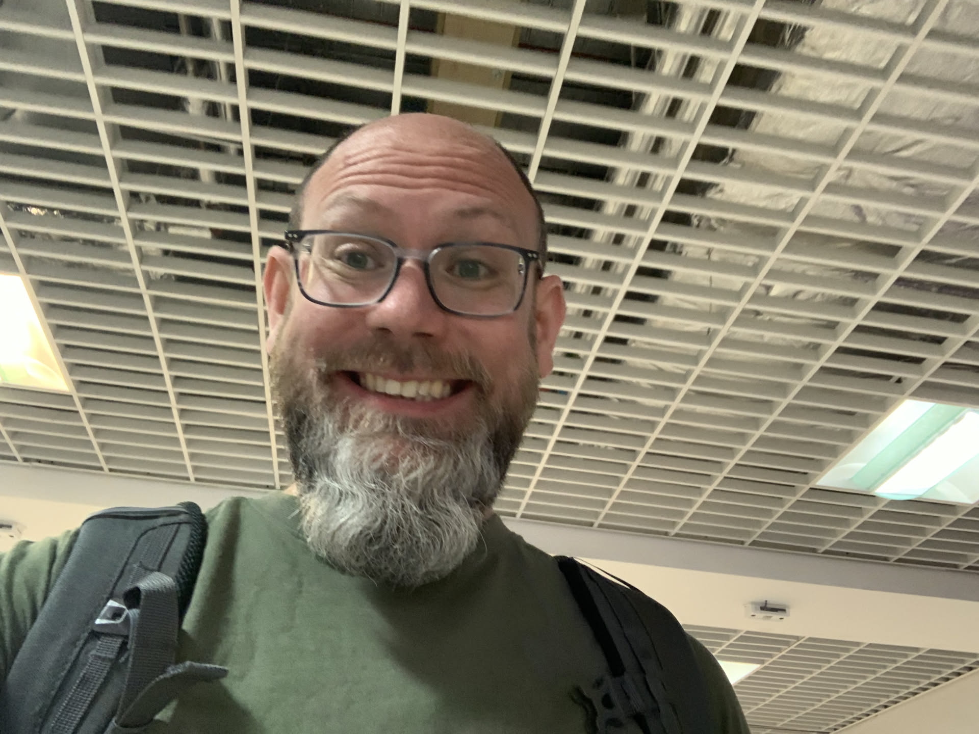 Stuart Thomas, a balding, bearded, spectacled, middle-aged white man. Grey and white hair, blue eyes, and a big grin on his face. Wearing a green t-shite and a rucksack.