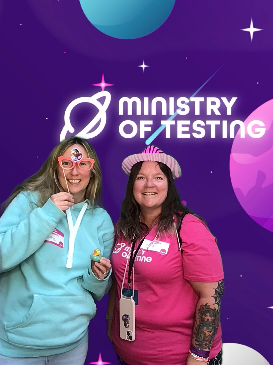 Two attendees pose together in a photobooth. 

On the left, attendee one is wearing a light blue hoodie that almost blends in with one of the planets in the background. They have blonde hair, are wearing playful glasses, and are smiling broadly. In their hand, they hold a small rubber duck that matches their hoodie. 

Attendee two, on the right, is dressed in a pink "Ministry of Testing" t-shirt with a matching pink zebra-striped silly hat. They are also smiling widely and looking at the camera. Their left arm, which is angled toward the camera, displays artistic tattoos. They wear a phone around their neck, along with a lanyard. 

Background: The green screen shows a fun outer space illustration. The primary color is purple, with cute stars and planets in pink, blue, and white. About a third of the way down, a logo appears. It reads "Ministry of Testing" in bold white text, with a blue shooting star and a white line drawing of Saturn.