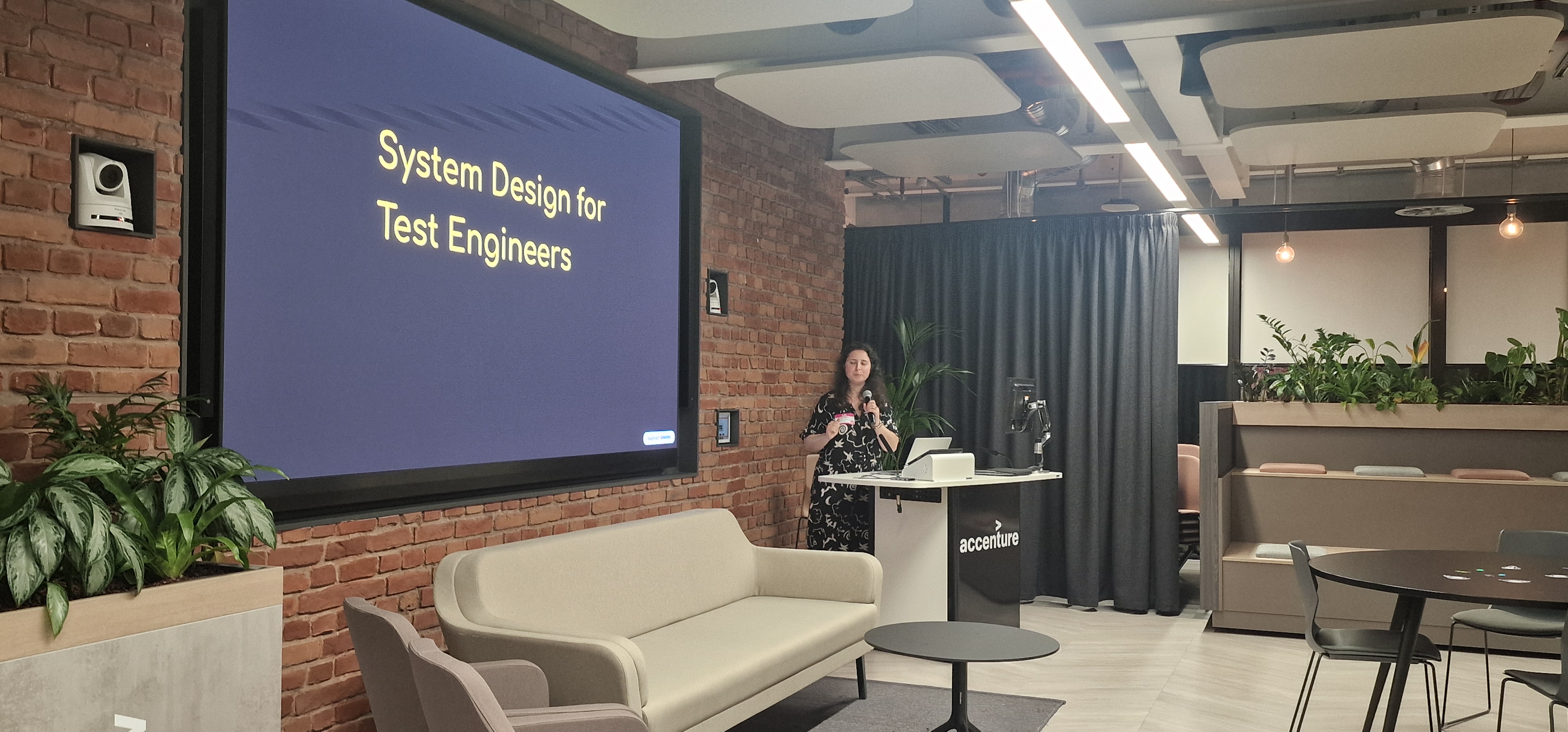A speaker stands behind a white Accenture-branded podium in a modern event space, holding a microphone. To their left... image