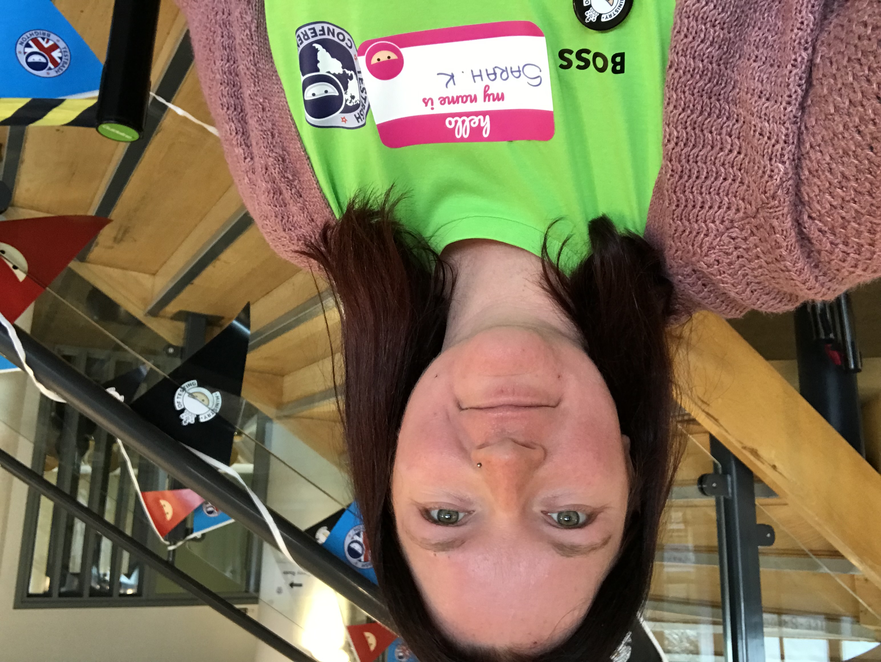 A selfie of Sarah K at her first TestBash in Brighton in 2019. She is wearing a shocking bright green MoT shirt standing by the main staircase in the venue that has MoT flags hung along the bannister.