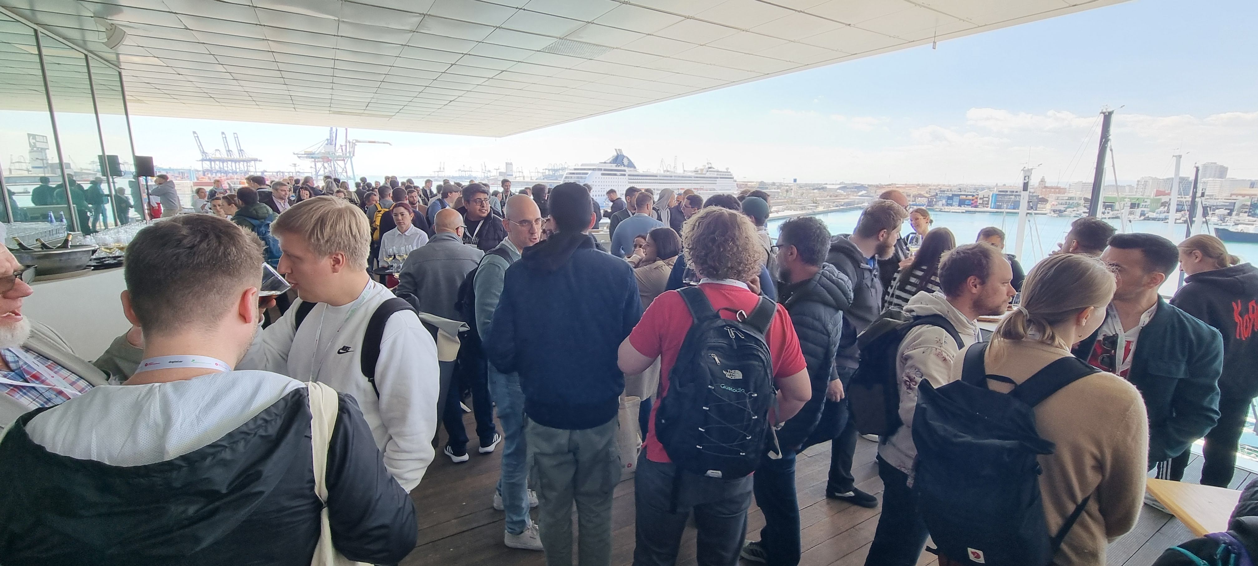 Lots of testers drinking something and talking with each other nearby a port, in a relaxed fashion, and a sunny day