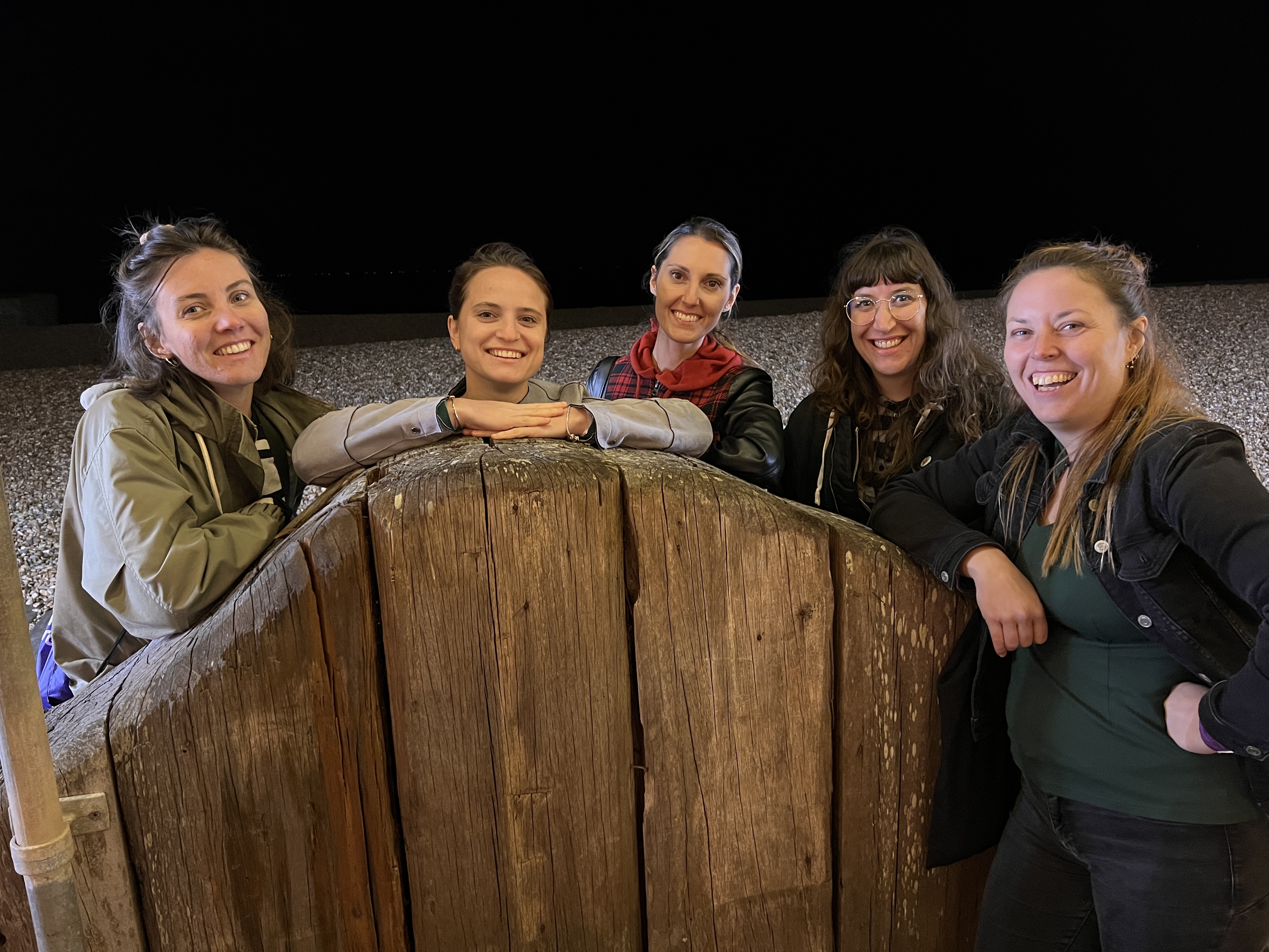 
A group of five smiling women stands behind a large, rustic wooden structure at night. The women are looking towards the camera. The woman on the far right rests her hand on the wooden structure.