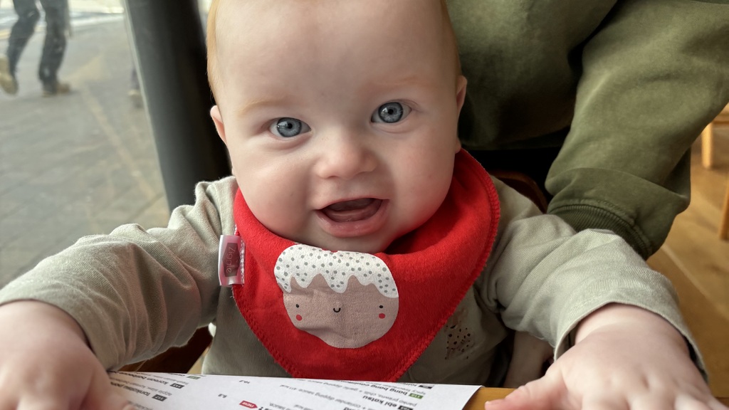 Harris looking at the camera while sitting at a café table, wearing a bright red bib, with an adult behind him. image