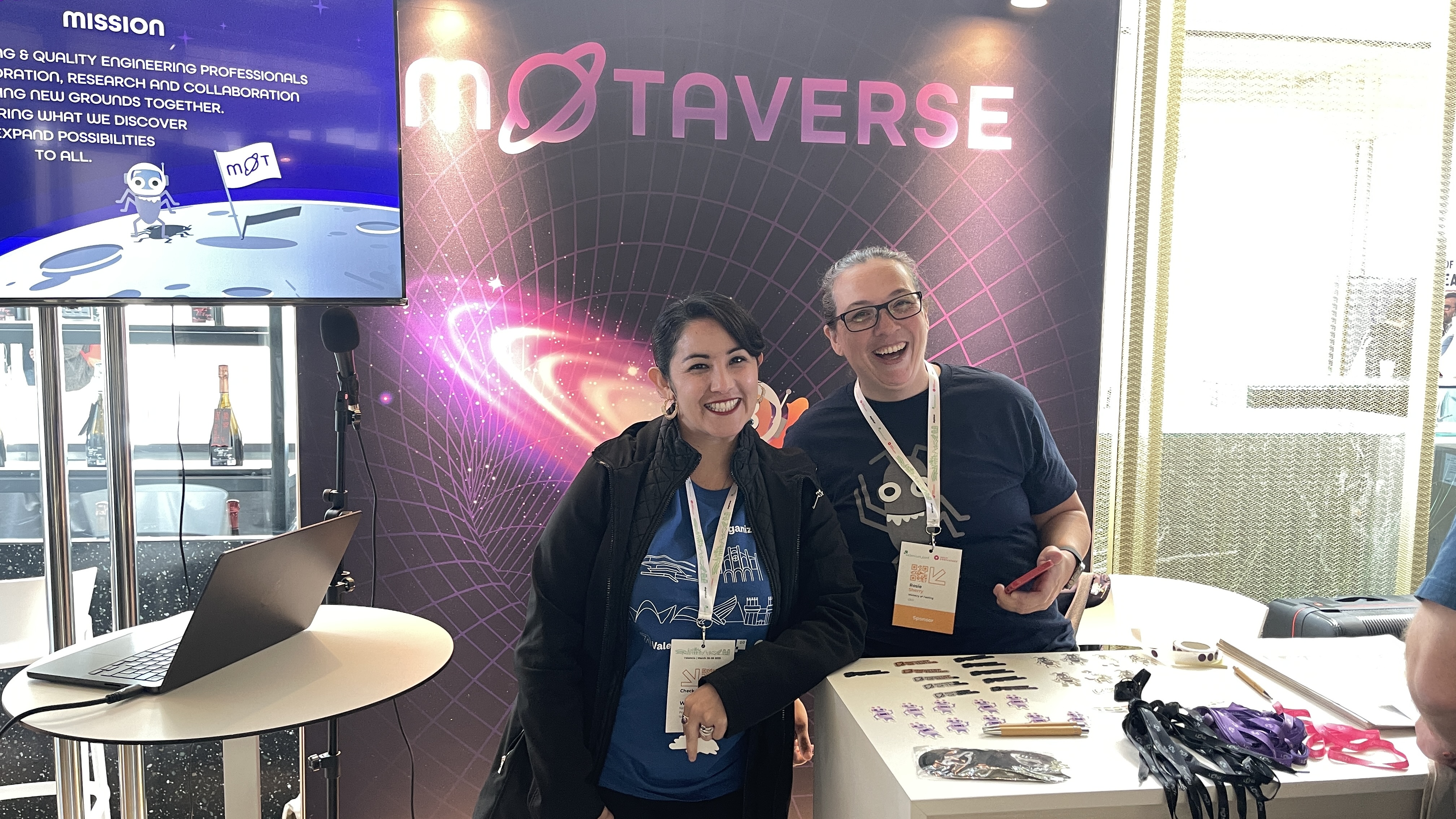 Rosie Sherry and Blanca J. Moreno Gallegos stand smiling at the Ministry of Testing stand, officially opening the MoTaverse portal at Selenium Conf 2025. Behind them is the MoTaverse galaxy-themed banner, with conference lanyards and stickers on the desk.