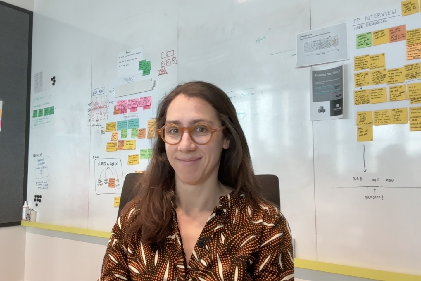 Some brown-haired white lady with a noisy top and a properly stickie note covered office wall behind her. 