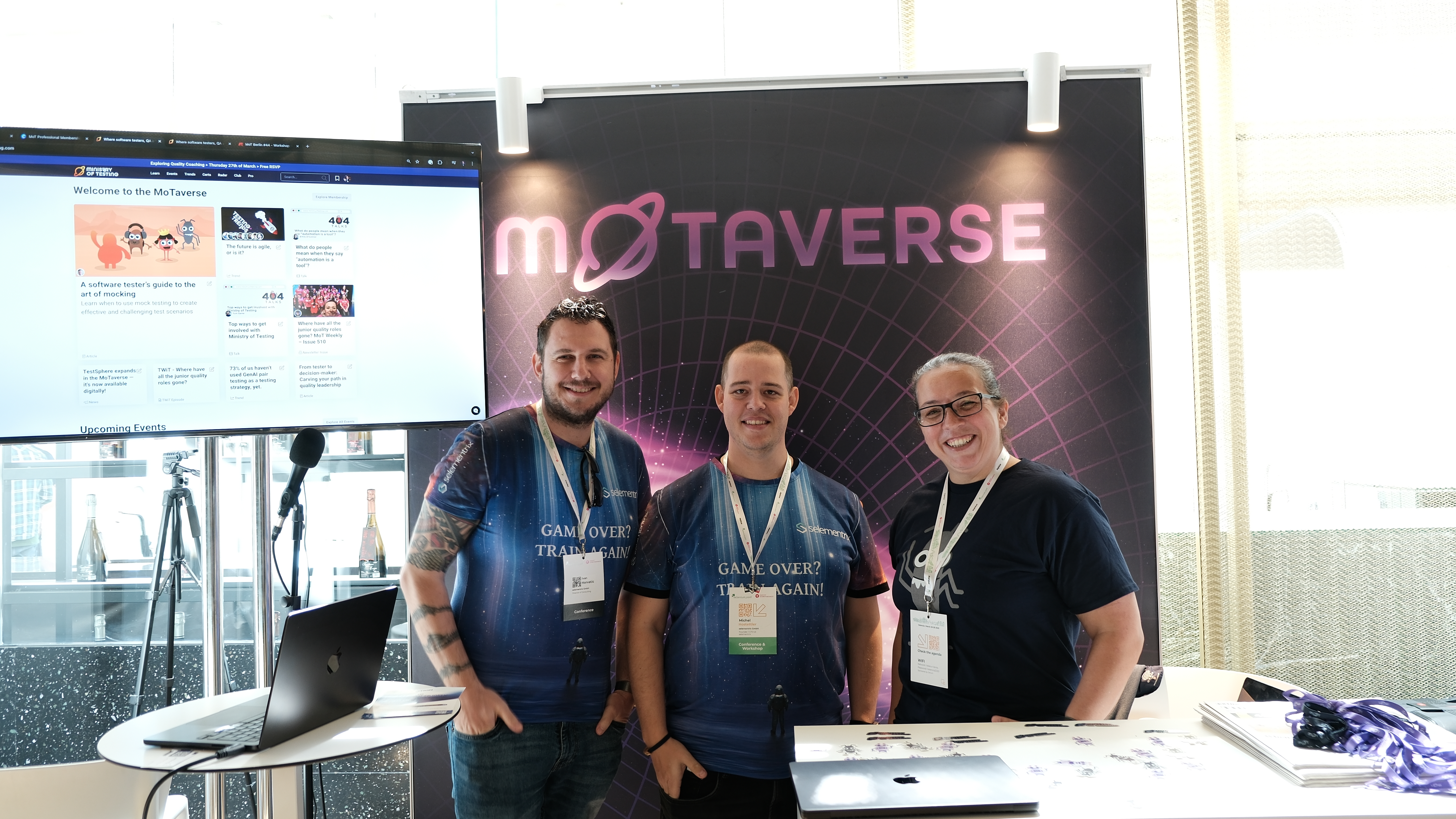 Ivan and Michel from Selementrix stand smiling with Rosie Sherry at the Ministry of Testing stand at Selenium Conf 2025, with the MoTaverse banner and a screen showing MoT content in the background.
