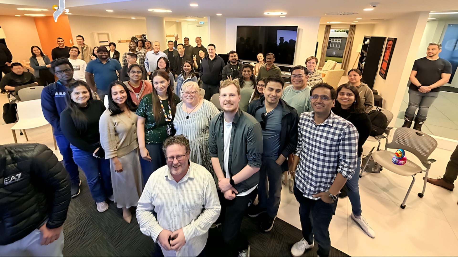 A diverse group of professionals stands in a modern, bright office space for a group photo. Approximately 30 to 40 people are present, most of whom are smiling and facing the camera. The attendees are dressed in business casual attire. The room features a large, wall-mounted monitor and an open area leading to another room with comfortable seating. A Bug, a duck and a seagull character have been added to the photo. 