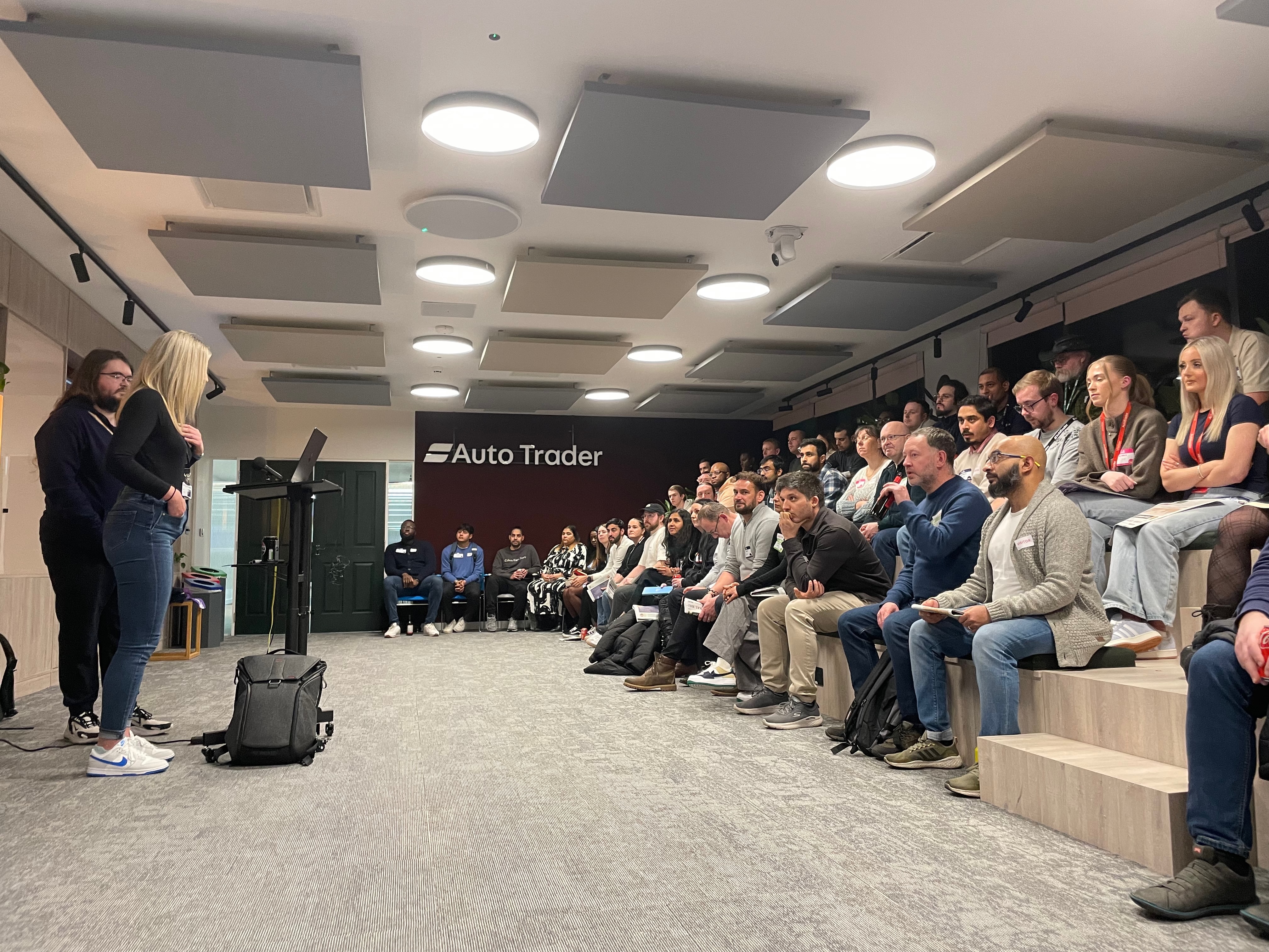 A photo of two speakers and an audience of about 60 people attending the meetup. There is an Autotrader wall sign in the background.