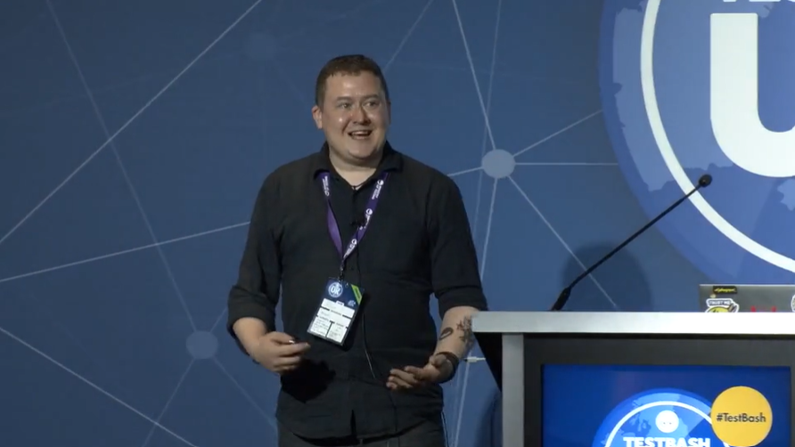 White male with shiny face dressed in black stood on the stage at TestBash UK 2023.