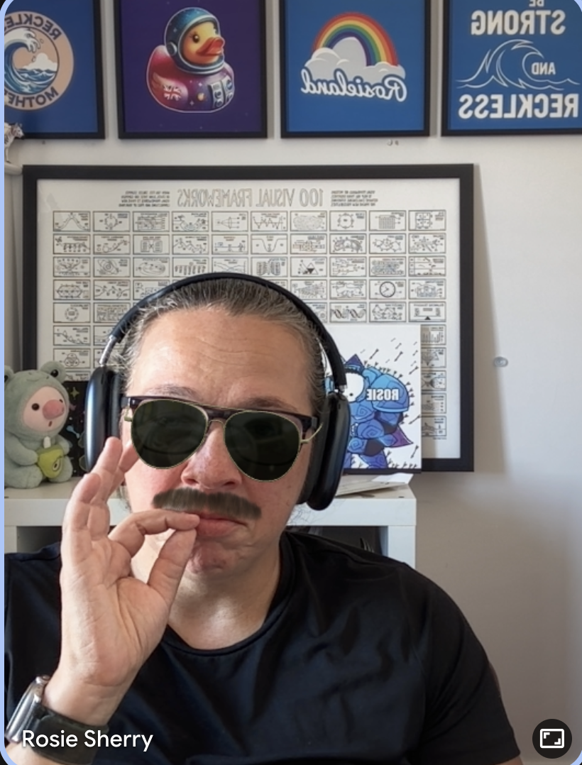 A Rosie screenshot from a virtual meeting, with sunglasses and a moustache. Her fingers are pinched, pretending she i... image