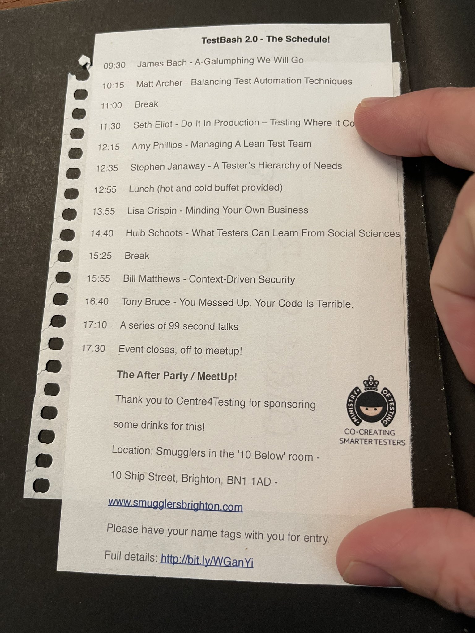 A printed schedule for "TestBash 2.0" is shown on a piece of perforated notebook paper. A person's fingers are holding the paper on the right and bottom edges. The schedule lists the event's talks and breaks from 09:30 to 17:30, followed by after-party details. Talks include titles such as “A-Galumphing We Will Go,” “Balancing Test Automation Techniques,” “Do It In Production – Testing Where It Counts,” and “You Messed Up. Your Code Is Terrible.” The after-party is at “Smugglers in the ‘10 Below’ room” at 10 Ship Street, Brighton. The Ministry of Testing logo appears at the bottom right.