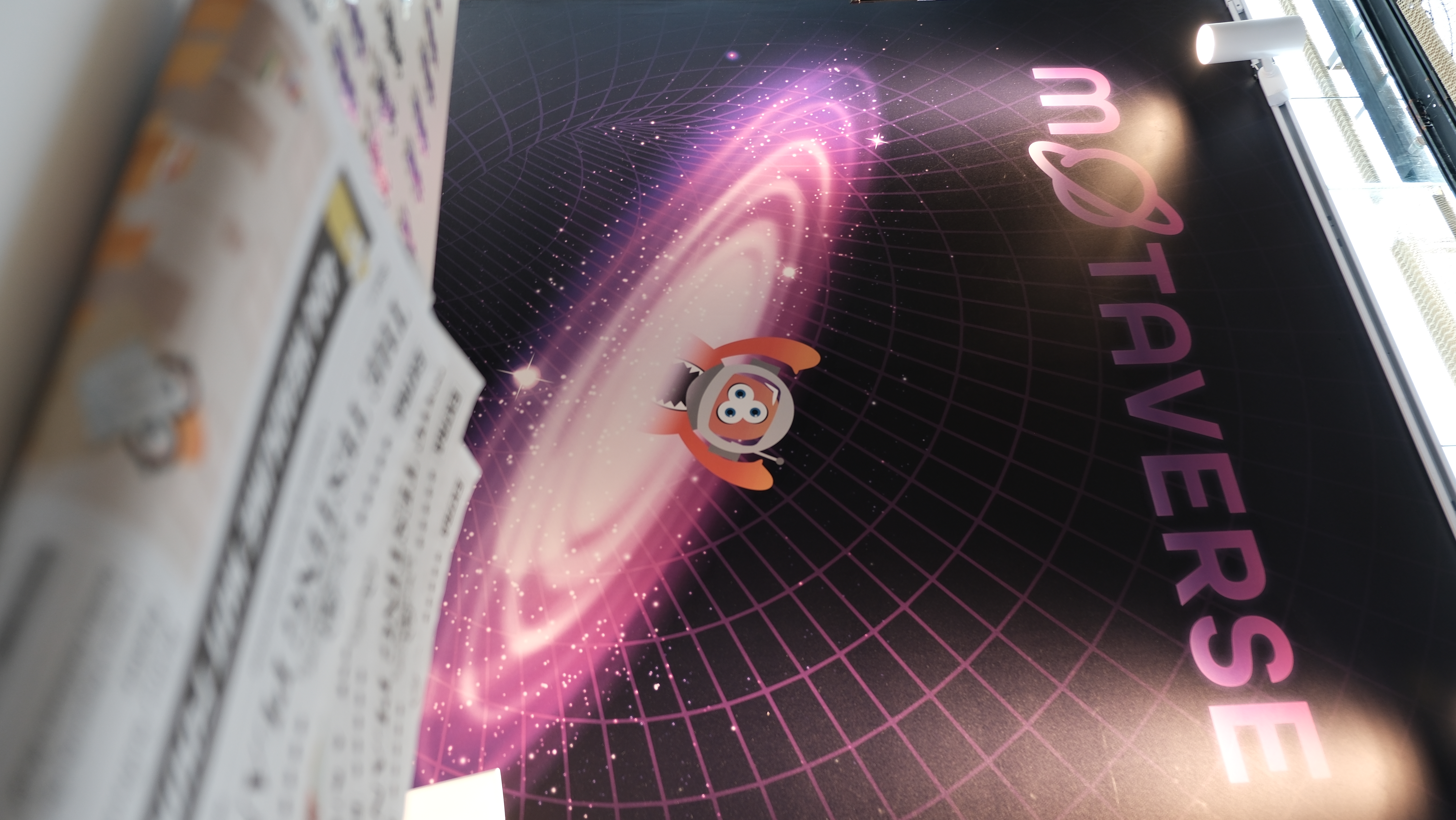 Backdrop of the Ministry of Testing's MoTaverse stand at Selenium Conf 2025, featuring a cartoon space-themed character flying through a glowing galaxy, with stickers and zines visible in the foreground.
